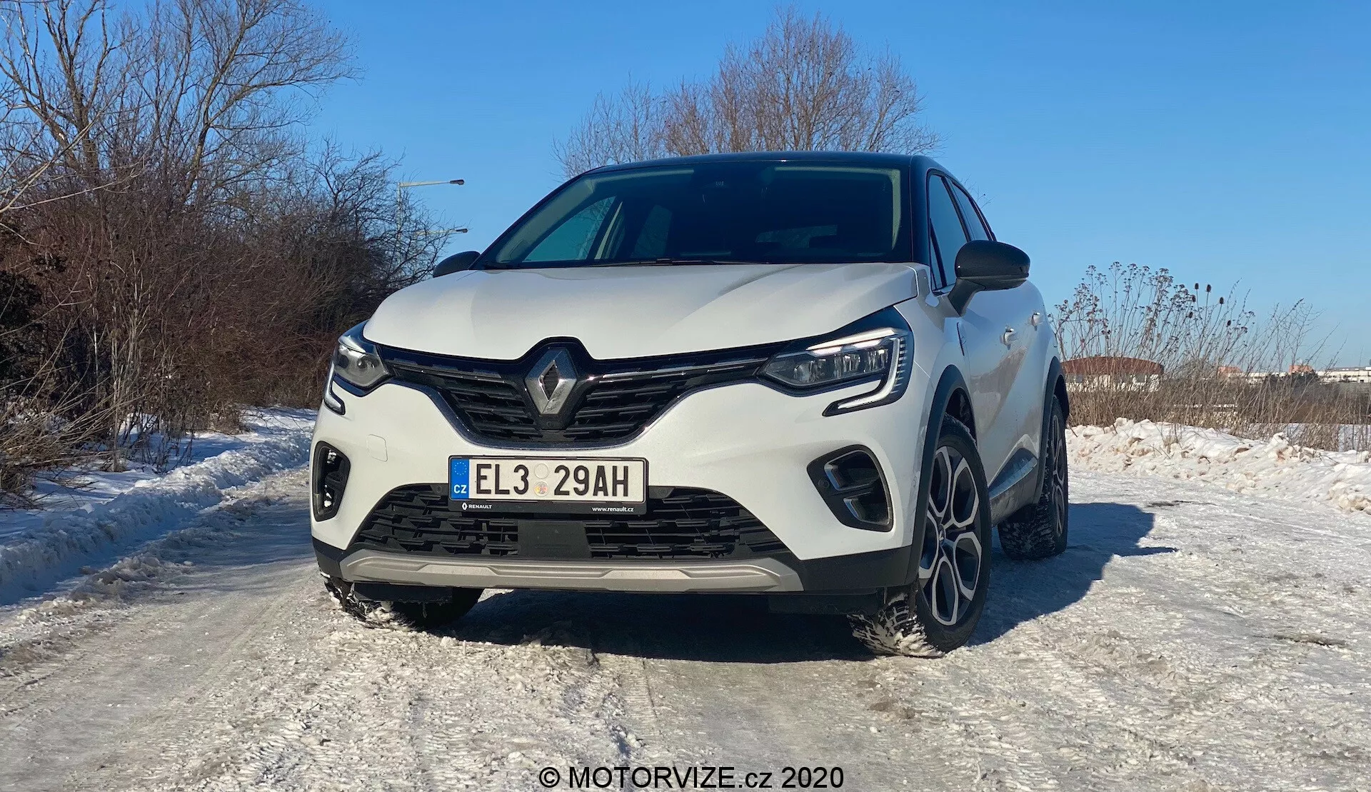 Vista frontal de um Renault Captur branco de 2020, fotografado num ângulo que favorece ligeiramente o lado esquerdo. O veículo moderno exibe um design distinto de grelha frontal Renault com um grande logotipo central, linhas angulares afiadas e contornos esculpidos. A grelha frontal completa, os faróis LED e as luzes diurnas angulares são claramente visíveis. O para-choques desportivo apresenta entradas de ar laterais proeminentes e uma seção central provavelmente para arrefecimento. O capô possui linhas elevadas que conferem um aspeto musculado. Parte das rodas dianteiras e das jantes de liga leve de múltiplos raios é visível na superfície revestida de neve, indicando condições de inverno. A matrícula europeia e a pintura brilhante que reflete a luz solar sugerem um dia ensolarado e claro. Sistemas avançados de assistência ao motorista, iluminação LED e motorização híbrida/elétrica são sugeridas pela letra 'E' na matrícula.