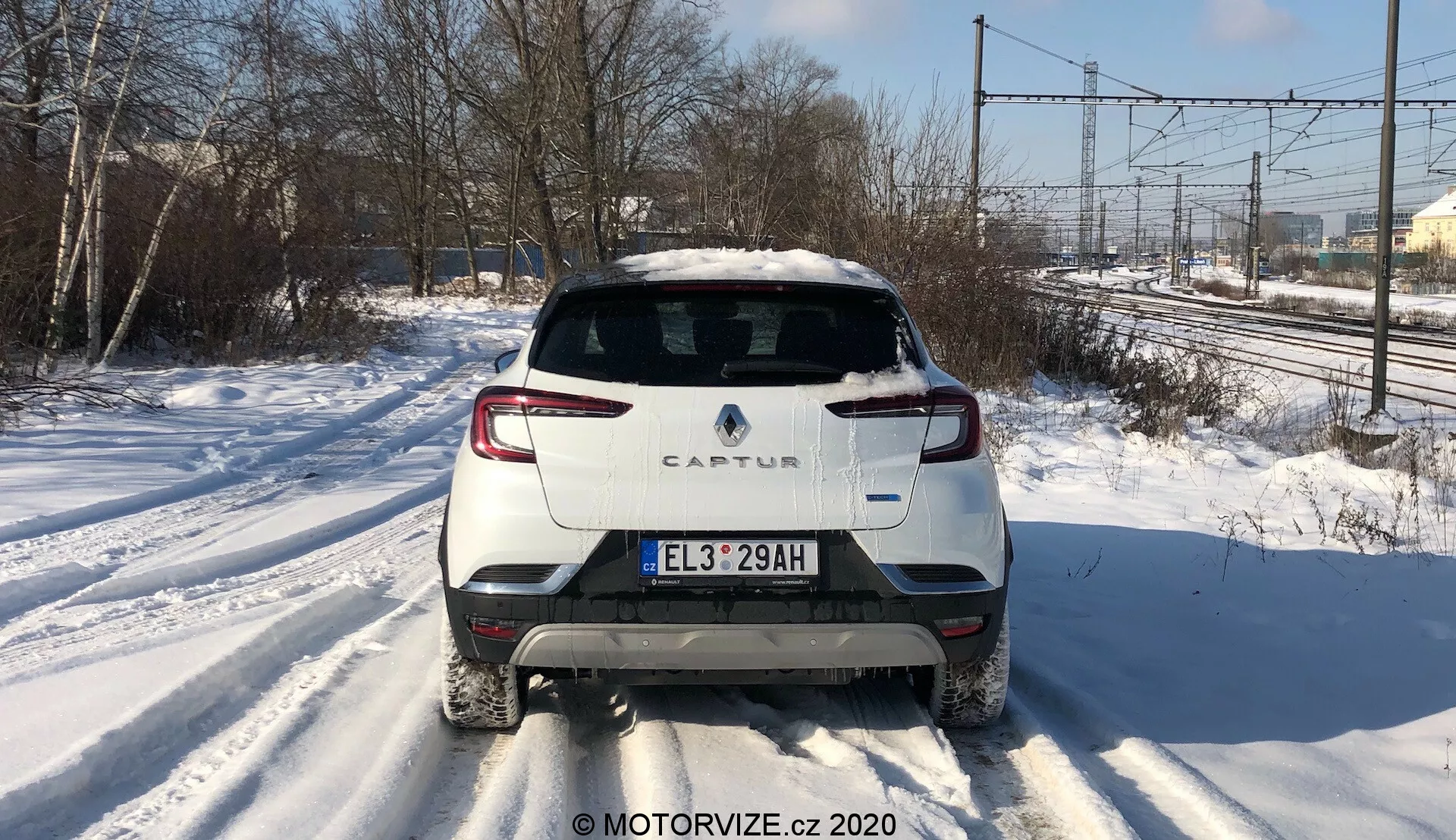Vista traseira centralizada do Renault Captur 2020 em uma estrada coberta de neve. A imagem destaca o design traseiro distinto, incluindo o esquema de cores de dois tons com carroceria branca e teto contrastante, iluminação traseira moderna em LED, placa de licença estilo europeu, limpador de para-brisa traseiro e um único tubo de escape do lado direito. A foto captura linhas de energia e árvores ao fundo sob luz do dia, sugerindo um contexto suburbano ou semi-rural.