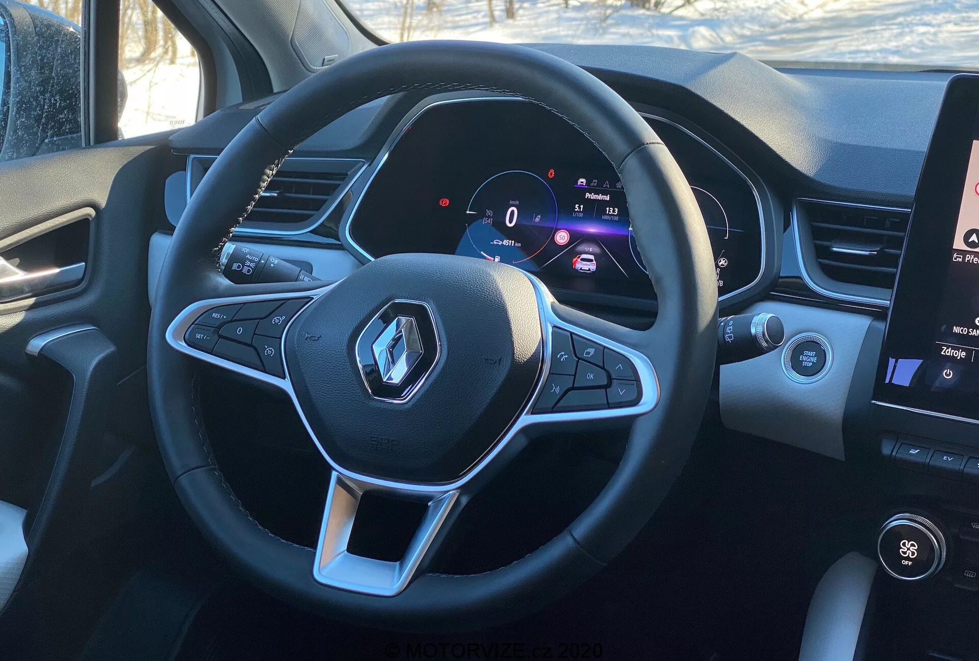 Vista interior do Renault Captur 2020 a partir da perspectiva do motorista, destacando o volante multifuncional esportivo de fundo plano com o logo da Renault e botões de controle para funções de áudio e controle de cruzeiro. O painel de instrumentos digital exibe uma velocidade estática de 0 km/h ao lado de um tacômetro, indicando tecnologia moderna e informações personalizáveis ​​para o motorista. À direita do volante, uma seção do sistema de infotainment com tela sensível ao toque vertical está visível, mostrando controles de temperatura e informações de mídia, em condições de luz do dia, oferecendo uma visão realista do assento do motorista.