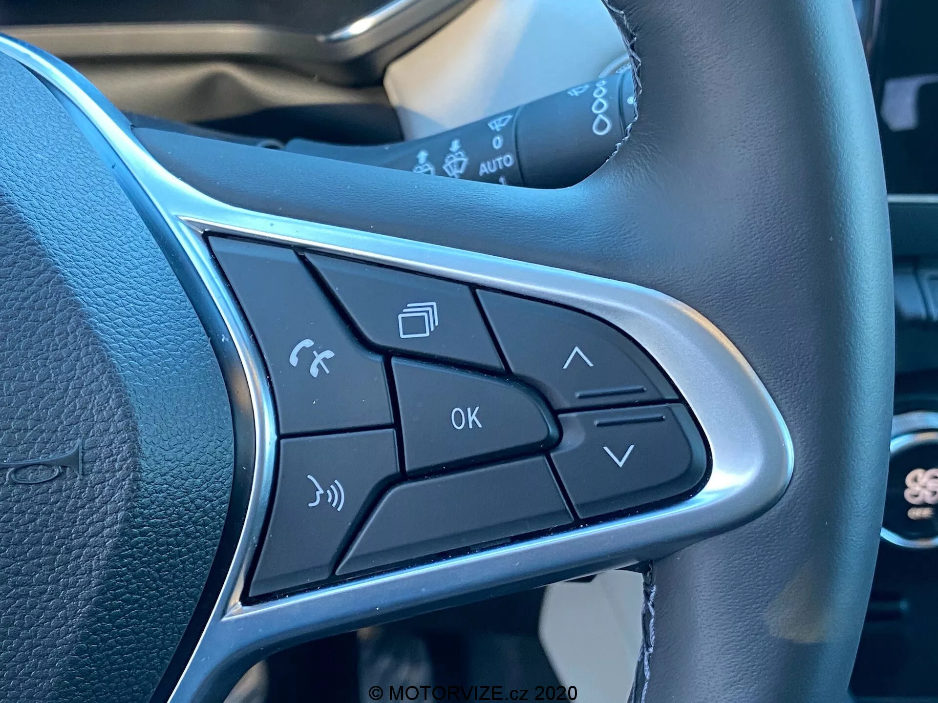Close-up dos botões de controle na haste do volante do Renault Captur 2020. Inclui ativação de comando de voz, gestão de chamadas telefônicas, controles de volume, botão OK e setas de navegação de menu. O design elegante inclui um acabamento prateado que destaca os botões, enfatizando a operação viva-voz e a conveniência.