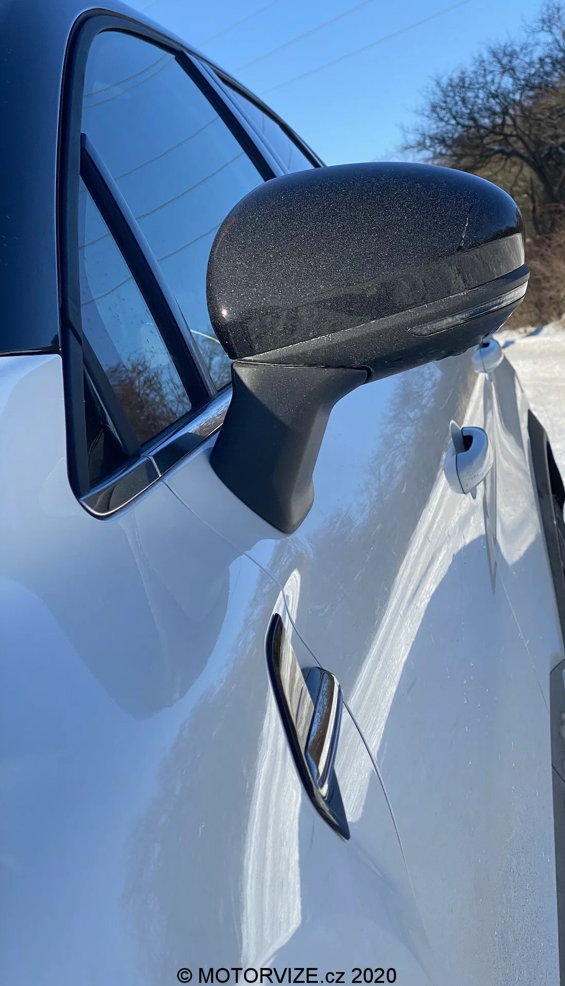 Vista detalhada em close do retrovisor, porta e painel frontal do Renault Captur 2020 branco. A foto, tirada de lado, logo à frente do retrovisor em um ângulo ascendente, destaca o design moderno e aerodinâmico do retrovisor com uma seção inferior texturizada preta para durabilidade. O suporte estilizado se integra perfeitamente ao quadro da porta. A maçaneta da porta embutida e a linha característica que a atravessa sugerem um design esportivo e elegante. Um vislumbre do farol e um reflexo do céu claro na janela realçam esta apresentação dinâmica.