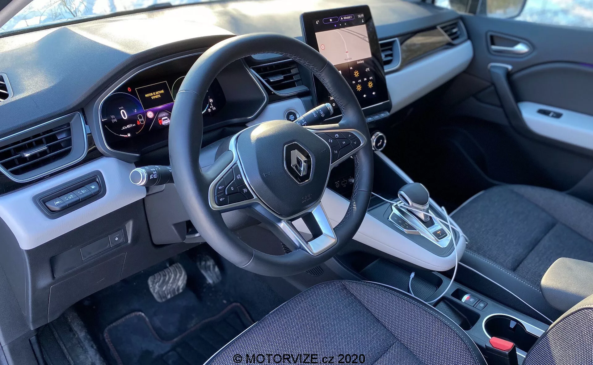 Visão interna de um Renault Captur 2020 a partir do banco traseiro, focando na área da cabine dianteira, incluindo o painel de instrumentos, volante com controles integrados, painel de instrumentos, console central com tela sensível ao toque e controles de clima, alavanca de câmbio automática e design moderno com uma mistura de elementos de tecido e plástico.