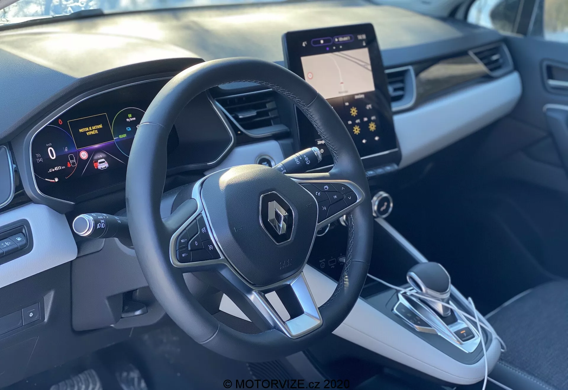 Vista interior do Renault Captur 2020 a partir do banco do passageiro dianteiro, apresentando o volante esportivo de três raios com emblema Renault e botões multifuncionais, painel de instrumentos digital com tela de alta resolução mostrando a velocidade e auxílios de navegação, e um console central elegante. O console central possui uma tela de infotainment moderna, alavanca de câmbio eletrônica e botão de freio de estacionamento eletrônico. O interior bicolor em cinza com design contornado e acabamentos proporciona uma sensação premium.