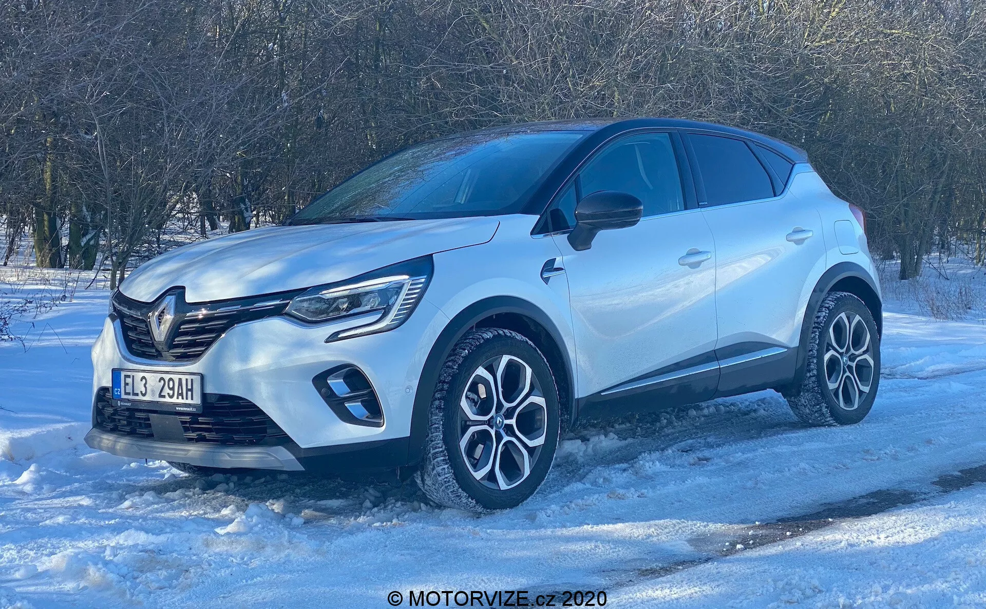 Vista de baixo ângulo de um SUV Renault Captur 2020 sobre uma superfície nevada, exibindo seu perfil frontal quartil. A luz do dia brilhante e o céu limpo destacam os elementos de design moderno, como a grade frontal proeminente com emblema, luzes diurnas em LED, rodas de liga leve complexas de duas cores, carroçaria esculpida com arcos de roda pronunciados, espelhos na cor da carroçaria com indicadores de direção integrados, janelas escurecidas para privacidade e maçanetas embutidas nas portas. A foto destaca a construção premium do veículo e suas potenciais capacidades de condução no inverno.
