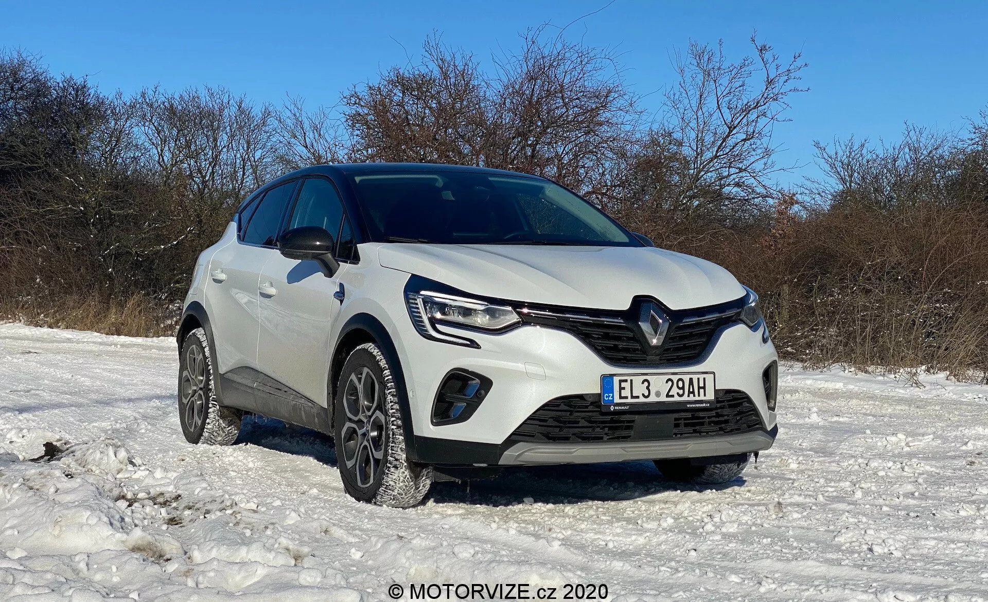 Renault Captur 2020 branco estacionado em terreno coberto de neve sob céu claro, mostrado de ângulo baixo de três quartos dianteiros exibindo a grade frontal, faróis com luzes diurnas de LED, luzes de neblina, para-choque dianteiro, pneus de inverno, logotipo Renault distinto, linhas esculpidas no capô, altura elevada, possíveis sensores de assistência ao estacionamento, espelhos laterais e condição geral limpa e bem cuidada, com marca d'água MOTORVIZE.cz © 2020.