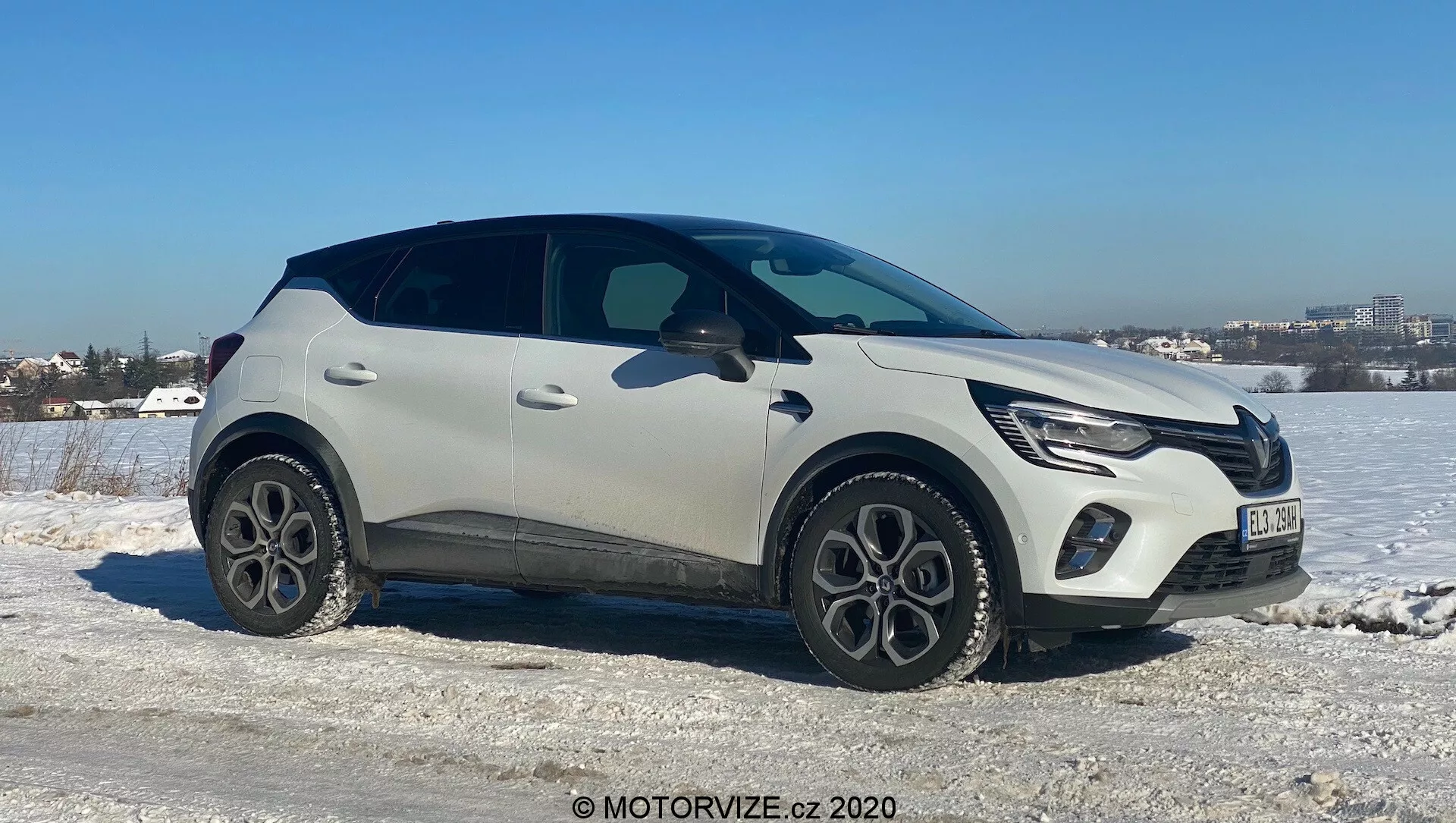 Renault Captur 2020 moderno SUV crossover posicionado em um terreno gelado e nevado com um céu claro ao fundo. O lado do passageiro está voltado para a câmera em um leve ângulo, mostrando a frente, o lado e parcialmente a parte superior do veículo. Características incluem luzes diurnas LED, grade frontal integrada com emblema do fabricante, pintura de carroceria em dois tons, claro e preto, rodas de liga leve de cinco raios, molduras protetoras de plástico, maçanetas de portas elegantes e embutidas e uma linha de janela afilada com um spoiler traseiro. Sugere tecnologia avançada como iluminação LED e entrada sem chave, ambientada em uma paisagem de clima frio próximo a uma área urbana.