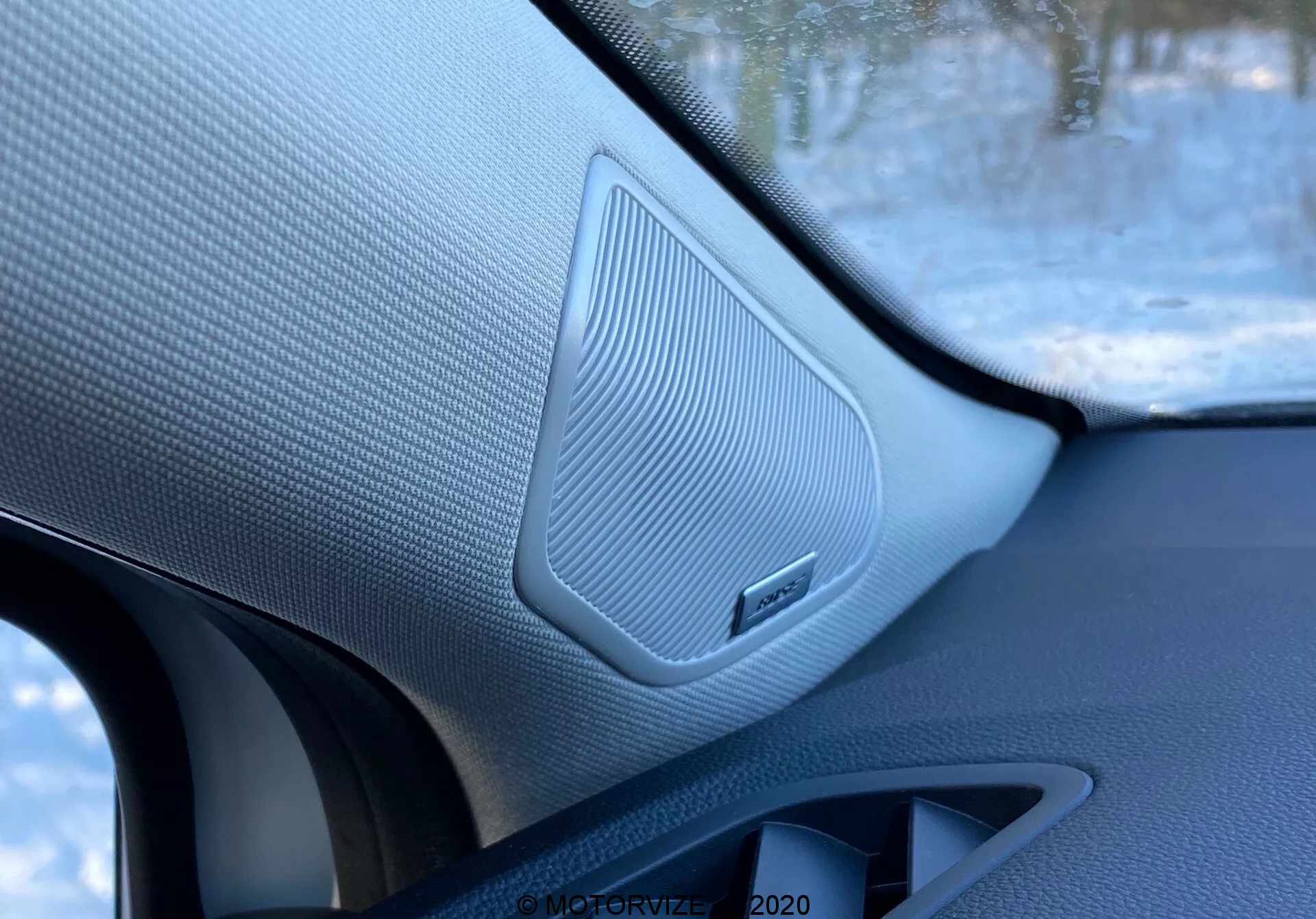 Imagem em close-up do interior de um Renault Captur 2020, destacando o alto-falante da porta dianteira do passageiro com uma grade texturizada em malha, rodeado por materiais contrastantes e um emblema da marca, tirada de dentro do veículo.