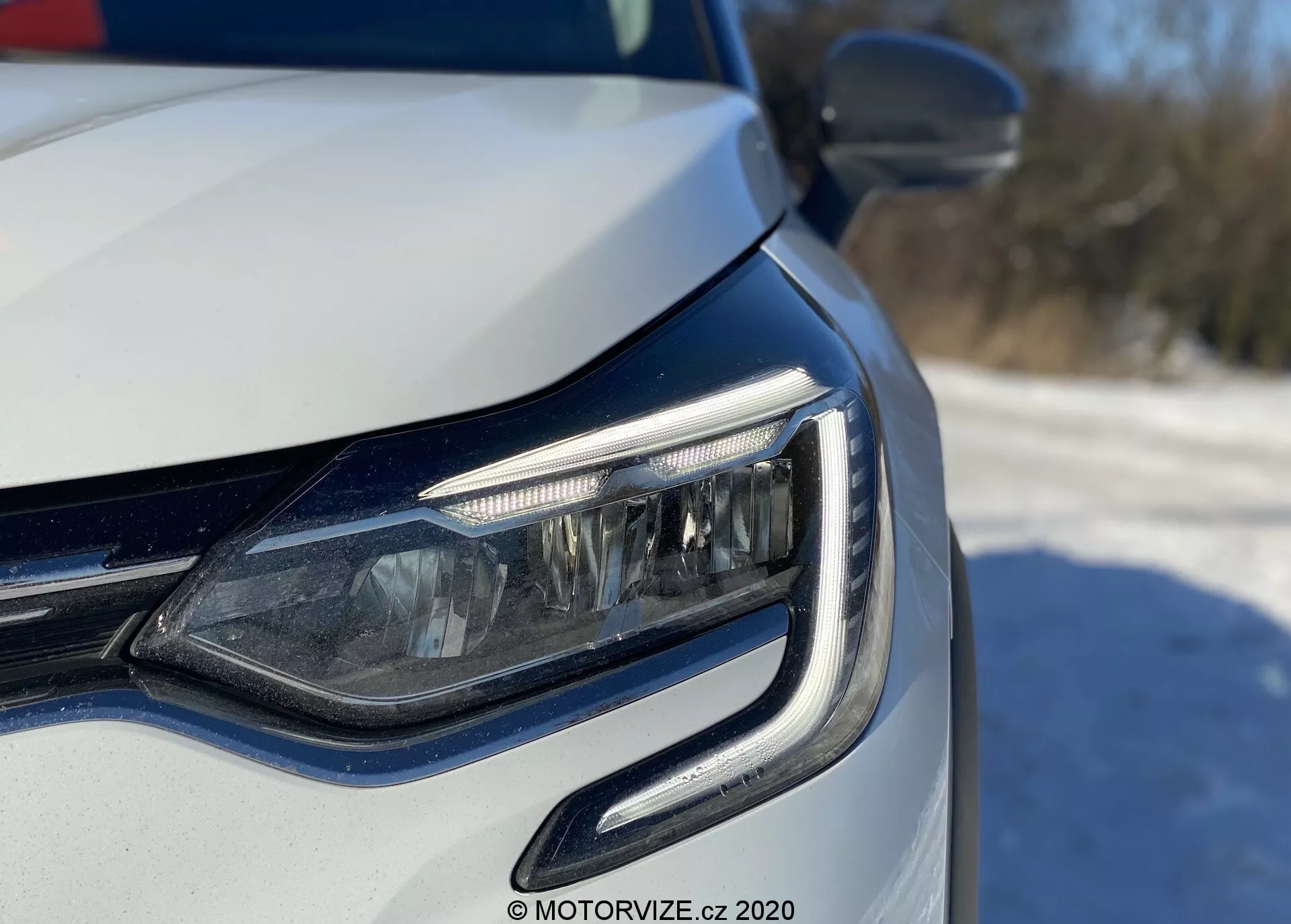 Close-up do canto frontal esquerdo de um carro moderno, focando no farol de LED e na grade, tirado de um ângulo baixo próximo ao nível do farol. As luzes de circulação diurna (DRL) apresentam um design curvo envolvendo a unidade do farol. Possíveis faróis adaptativos ou luzes de curva visíveis, com acabamento de pintura metálica e sinais de mudança de direção integrados no espelho retrovisor externo. Iluminação natural brilhante sugere um cenário externo diurno.