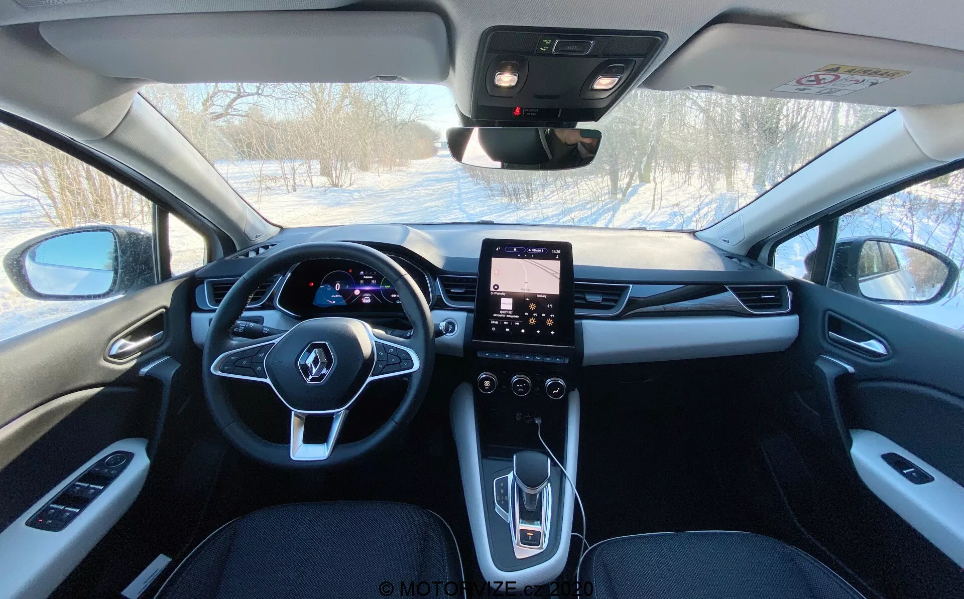 Vista interna de um Renault Captur 2020 da área dos assentos traseiros olhando para frente, mostrando seu estofado de cor escura, espelho retrovisor central, painel de instrumentos digital iluminado atrás do volante multifuncional, sistema de infotainment com tela sensível ao toque no console central e controles para janelas elétricas, espelhos laterais, controle de clima, e provavelmente para teto solar e botão de emergência SOS.