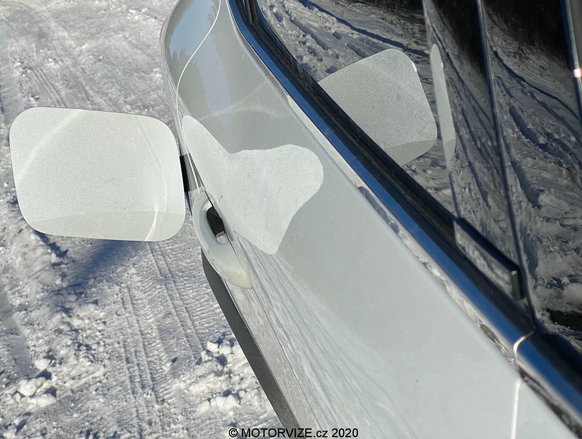 Vista lateral de close-up de um Renault Captur 2020 em um ambiente de neve ao ar livre. O foco está no retrovisor direito, dobrado para a frente, possivelmente indicando uma função de dobramento automático ou manual. A carcaça do espelho tem duas películas protetoras transparentes em forma de paralelogramo, provavelmente para proteção durante a entrega ou lavagem. O carro, em um tom escuro brilhante como preto ou azul escuro, parece bem conservado com reflexos que sugerem uma superfície limpa e polida. O solo ao redor e os lados do carro estão cobertos de neve. Parte da maçaneta da porta dianteira, com um botão de entrada sem chave ou sensor de toque, também é visível.