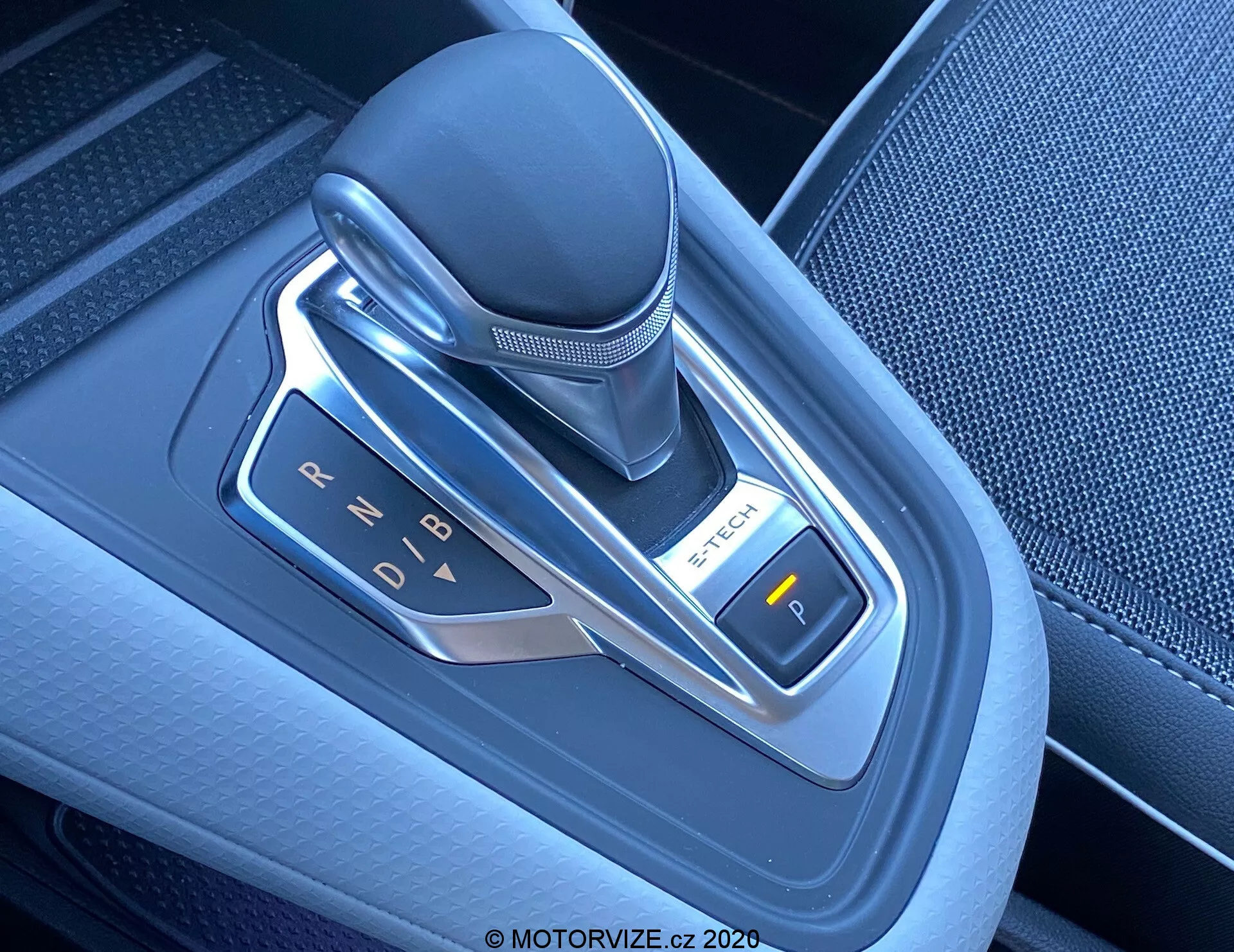 Imagem de close-up de uma alavanca de câmbio automática moderna em um Renault Captur 2020, localizada no console central. O seletor eletrônico de marchas está posicionado verticalmente e é feito de couro, plástico e detalhes metálicos. A fotografia, tirada de um ângulo superior ligeiramente à direita, destaca as indicações de marchas (P, R, N, D, B) iluminadas por luzes LED. À direita, é visível um botão rotulado com 'P', provavelmente para o freio de estacionamento eletrônico. A área ao redor da alavanca de câmbio apresenta plástico texturizado de alta qualidade com detalhes metálicos, refletindo a tecnologia avançada e o design contemporâneo do interior do veículo.