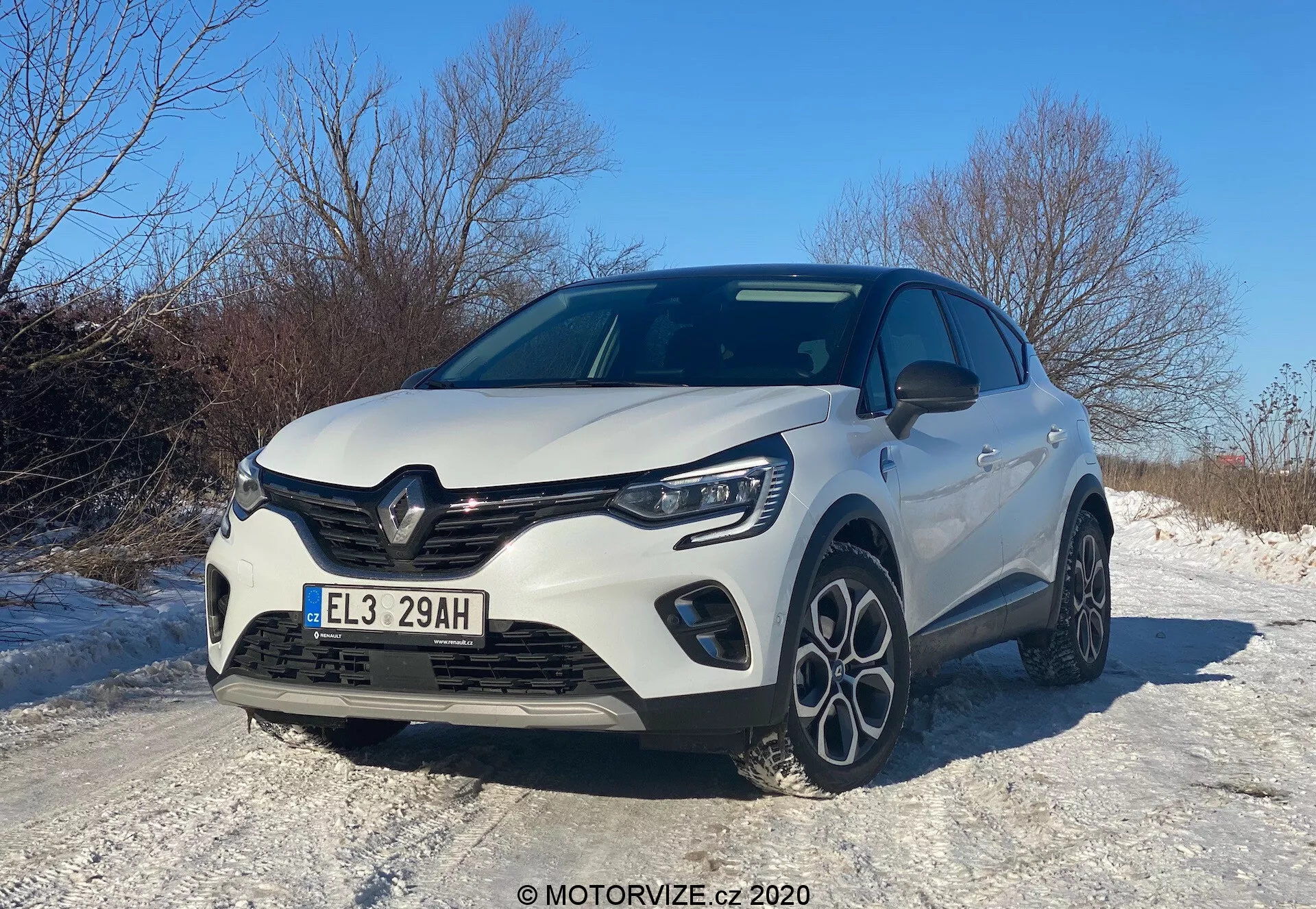 Veículo hatchback moderno Renault Captur 2020 capturado de um ângulo baixo de três quartos frontal, mostrando a grade frontal, capô, faróis de LED, para-choque dianteiro com luzes de nevoeiro integradas e um perfil lateral parcial. Os elementos de design incluem uma grade proeminente com o emblema do fabricante, linhas esculpidas no capô e rodas de liga leve de raios múltiplos, tudo sob um céu claro durante o dia, refletindo uma estética esportiva e aerodinâmica, possivelmente destacando uma versão aprimorada ou pacote esportivo. O carro está situado em uma estrada coberta de neve, sugerindo características adequadas para condução de inverno.