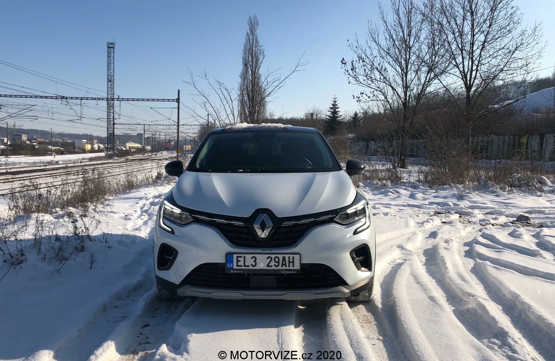 Vista frontal direta do Renault Captur 2020 em uma estrada coberta de neve, exibindo em detalhe a grade frontal, faróis, capô esculpido e para-choques dianteiro durante o dia. Rastros de pneus são visíveis, evidenciando movimento recente. O design do veículo sugere um estilo aerodinâmico, com emblema da marca destacado e sistema de iluminação avançado. Sensores ou câmeras de assistência ao motorista podem estar integrados no para-choques dianteiro.