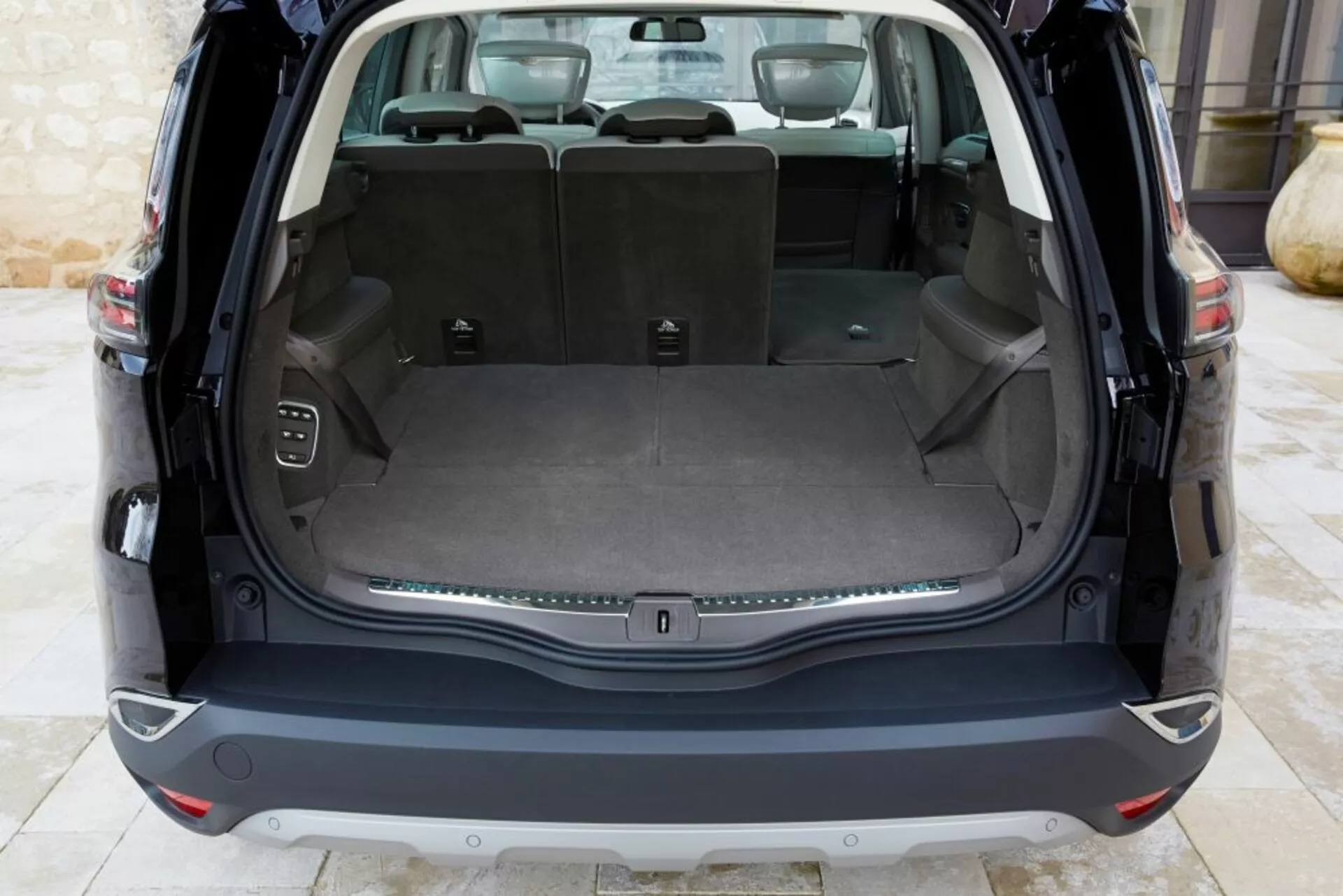 Vista interior traseira do Renault Espace 2015 mostrando uma área de carga limpa com o porta-malas aberto, apresentando um piso de carga plano, assentos traseiros rebatíveis 60/40, uma tomada de energia, ganchos de carga, botão de controle para o porta-malas eletrônico, e uma faixa decorativa cromada, sob iluminação natural.