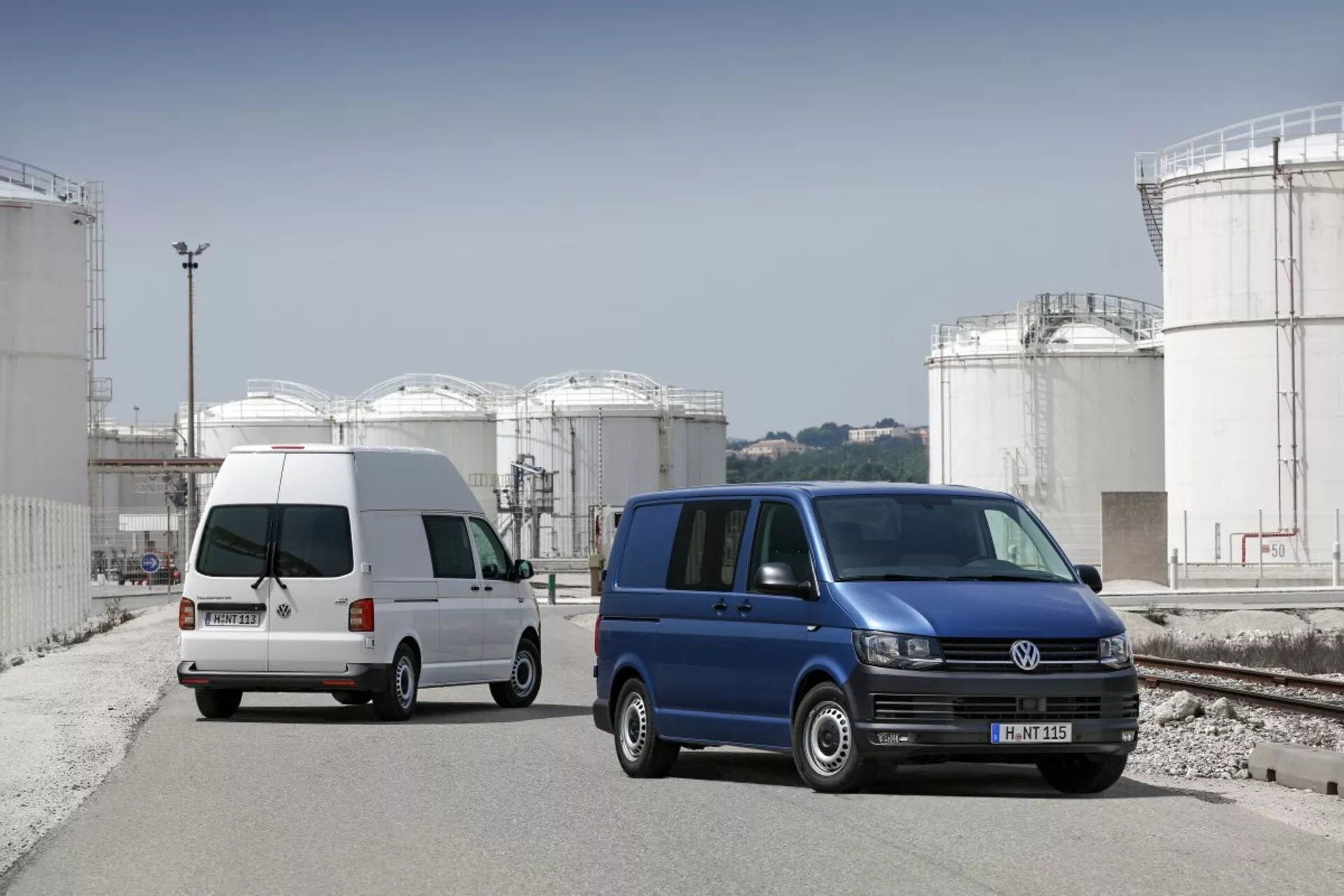 Duas vans Volkswagen Transporter estacionadas em um local industrial com grandes tanques de armazenamento ao fundo, exibindo seu design de utilidade comercial, portas laterais deslizantes e rodas de liga padrão.