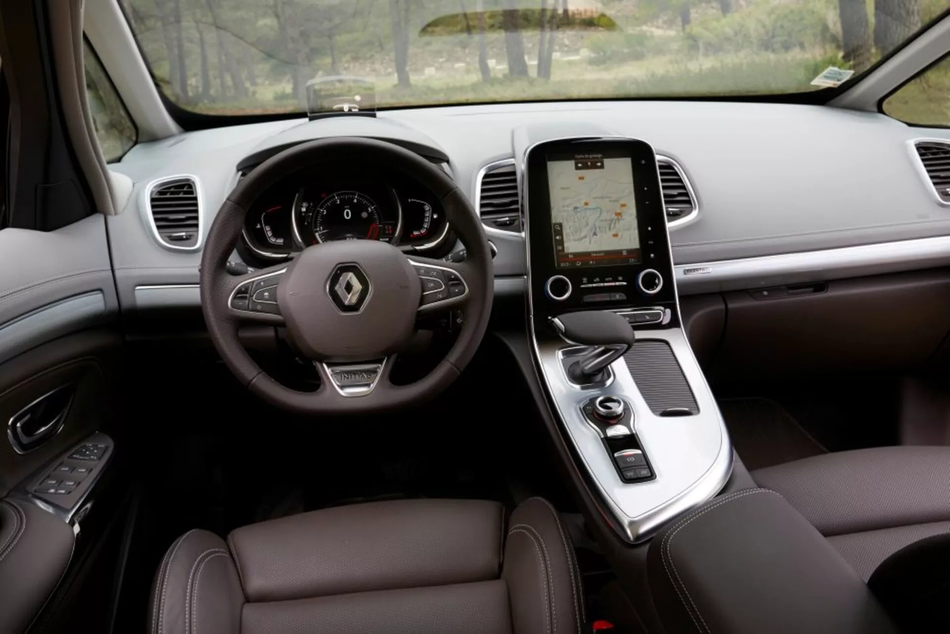 Vista interior do Renault Espace 2015 da perspectiva do assento do condutor, exibindo um design luxuoso, com volante de três raios com botões de controle, cluster de instrumentos digital, tela sensível ao toque vertical no console central para o sistema de infotainment, câmbio automático, assentos revestidos em couro com ajuste eletrônico, e saídas de ar no painel para controle do clima