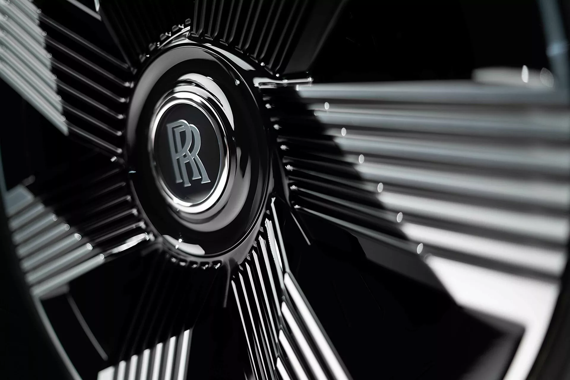 Detalhe do aro da roda do Rolls-Royce Spectre de primeira geração 2023 com o emblemático emblema RR em cromo contra um design escuro de múltiplos raios, destacado por fotografia com profundidade de campo focalizada