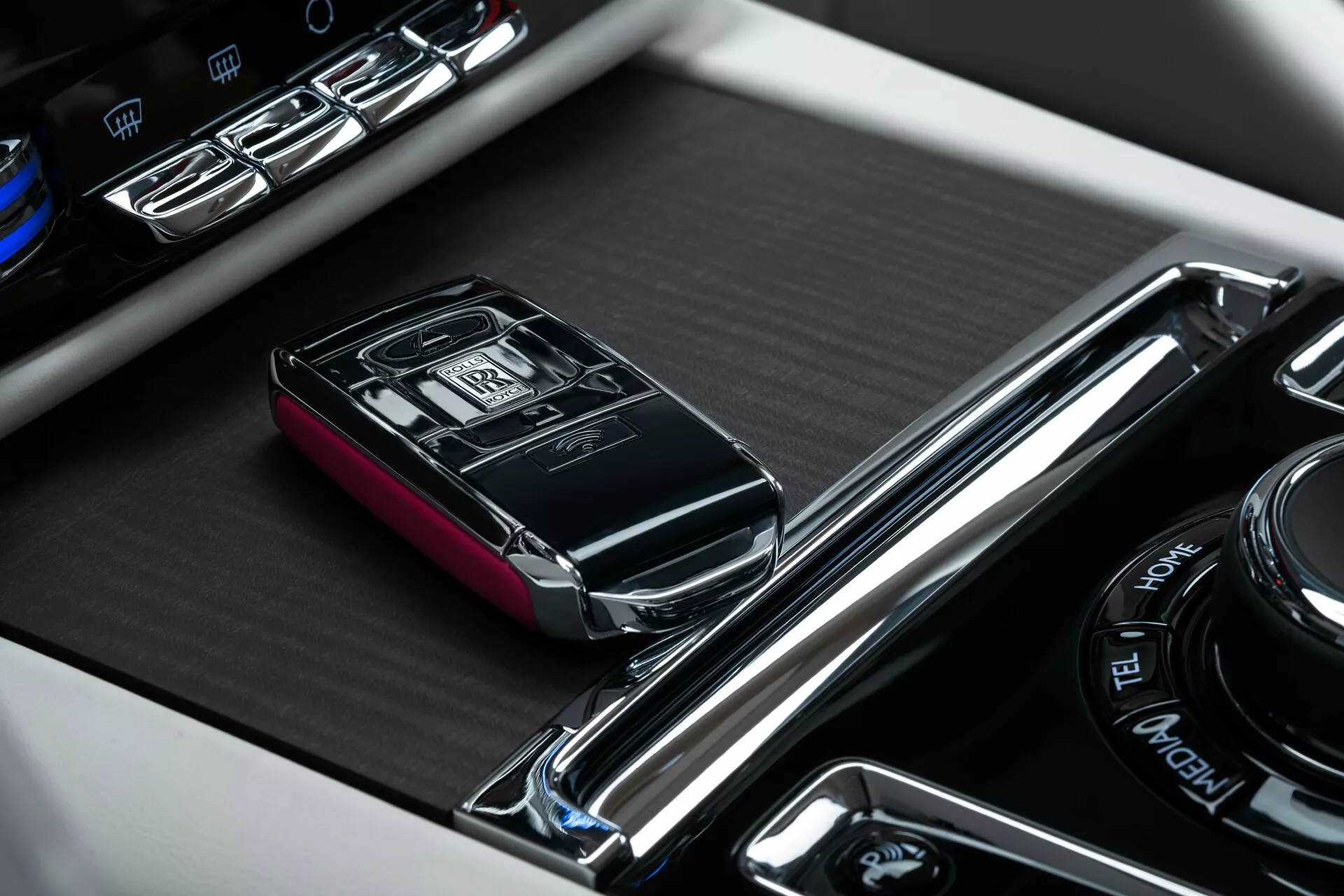 Close-up do console central do Rolls-Royce Spectre 2023 com foco no chaveiro de luxo do carro em seu slot designado, cercado por acabamento cromado e adjacente a botões de controle iluminados e um dial multimídia rotativo.