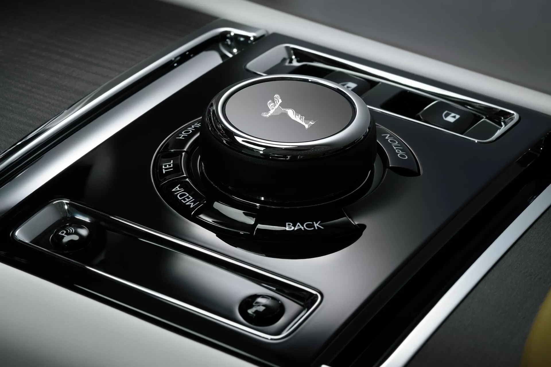 Vista de perto do console central do Rolls-Royce Spectre 2023 primeira geração, mostrando o botão de controle do infotainment com superfície sensível ao toque, cercado por botões de funções claramente etiquetados em um painel preto de alto brilho, situado em metal escovado para um design interior de luxo