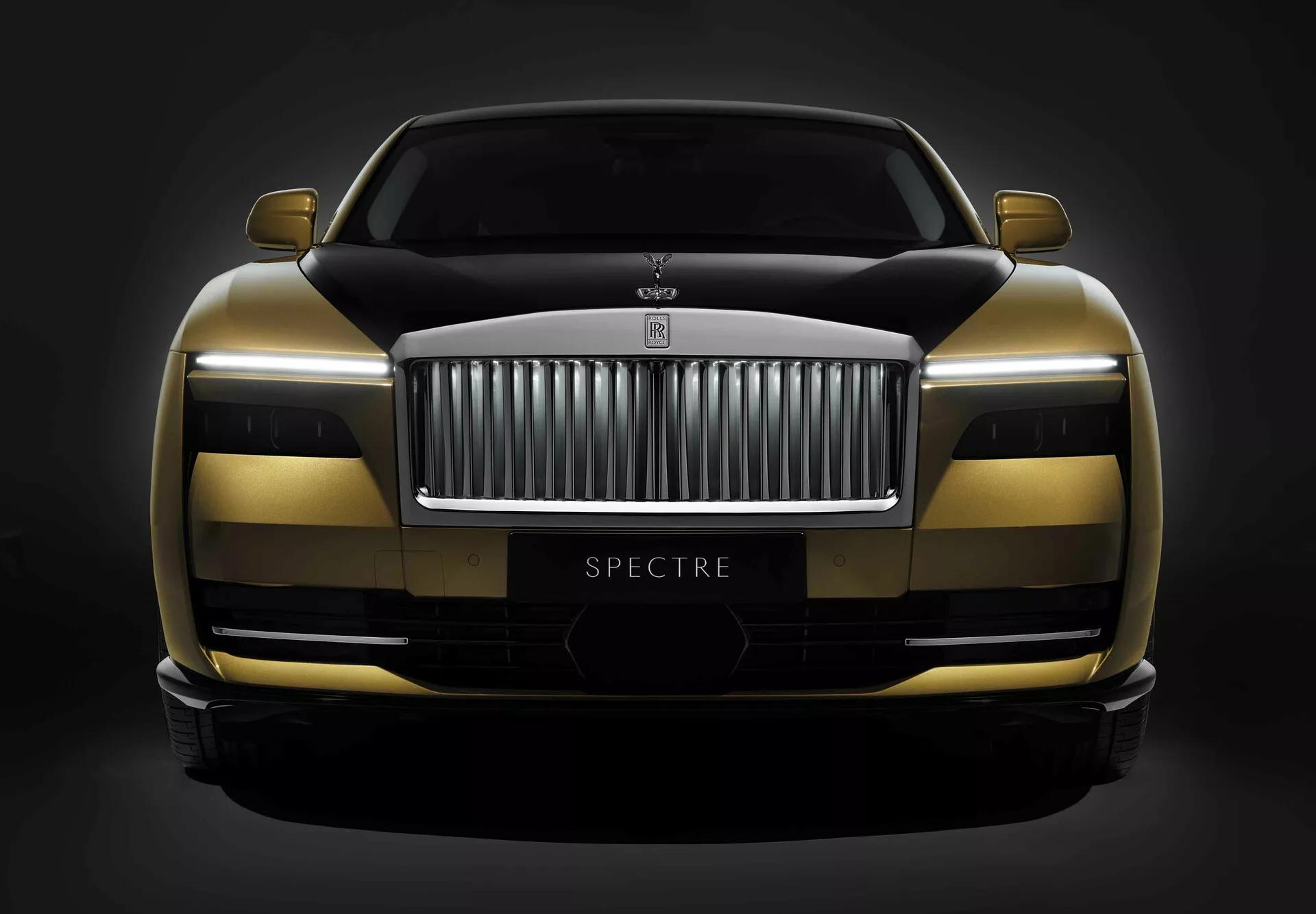 Rolls-Royce Spectre 1ª geração 2023 apresentando um esquema de pintura em dois tons, tecnologia avançada de faróis LED e um design aerodinâmico com o nome do modelo 'SPECTRE' visível, capturada de um ângulo frontal baixo contra um fundo escuro.