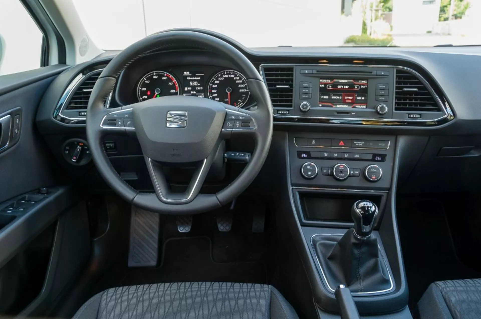 Vista interior de um Seat Leon SC de 2016 a partir da perspectiva do assento do passageiro frontal, mostrando o volante com o logotipo do fabricante e controles montados, cluster de instrumentos digital, sistema de infotainment, controles de clima, alavanca de câmbio manual e porta do motorista com opções de energia.