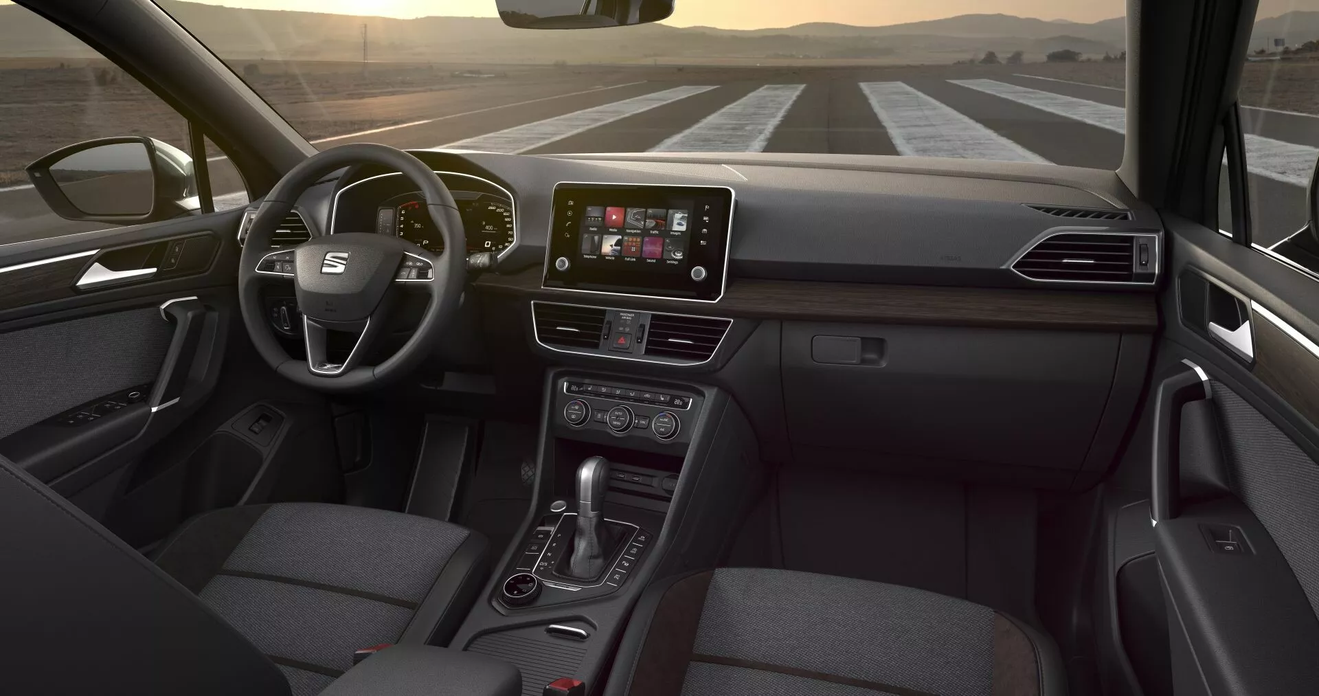 Perspectiva interior do Seat Tarraco 2019 a partir do assento traseiro, apresentando volante multifuncional, display central touchscreen de infotenimento, botões de controle de clima, alavanca de transmissão automática e assento do passageiro com estofamento em tecido, sob uma iluminação interior sutil
