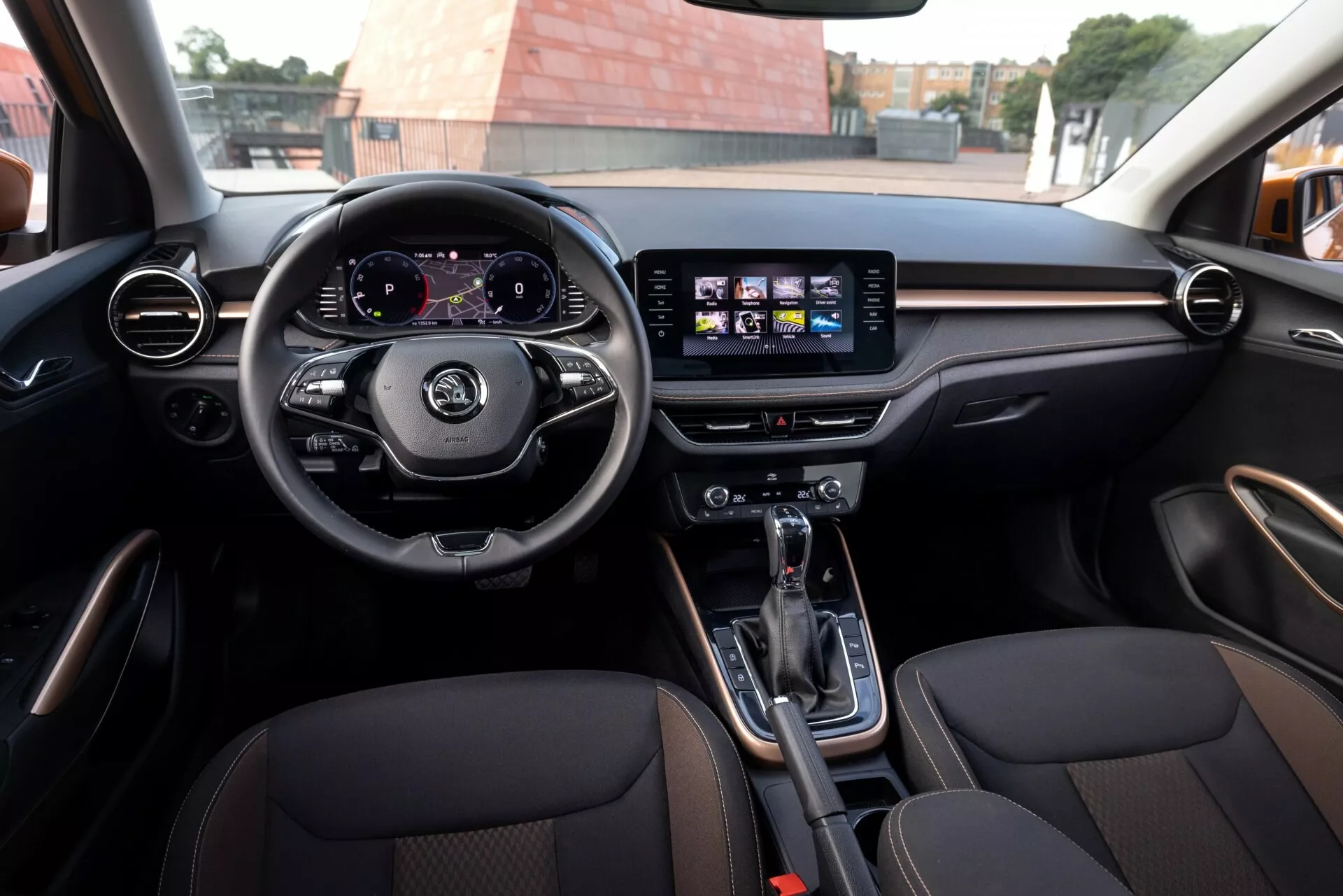 Vista interior do Skoda Fabia 2021 a partir do banco traseiro, mostrando um volante de três raios com controles, painel de instrumentos digital exibindo velocímetro e tacômetro, e sistema de infotainment central com tela sensível ao toque com ícones de navegação e música. Ventiladores circulares e horizontais com acabamento que imita madeira ou metal, transmissão automática com freio de estacionamento eletrônico no console central, assentos dianteiros esportivos revestidos em couro com costuras contrastantes. Ambiente moderno e espaçoso que indica luxo e esportividade.