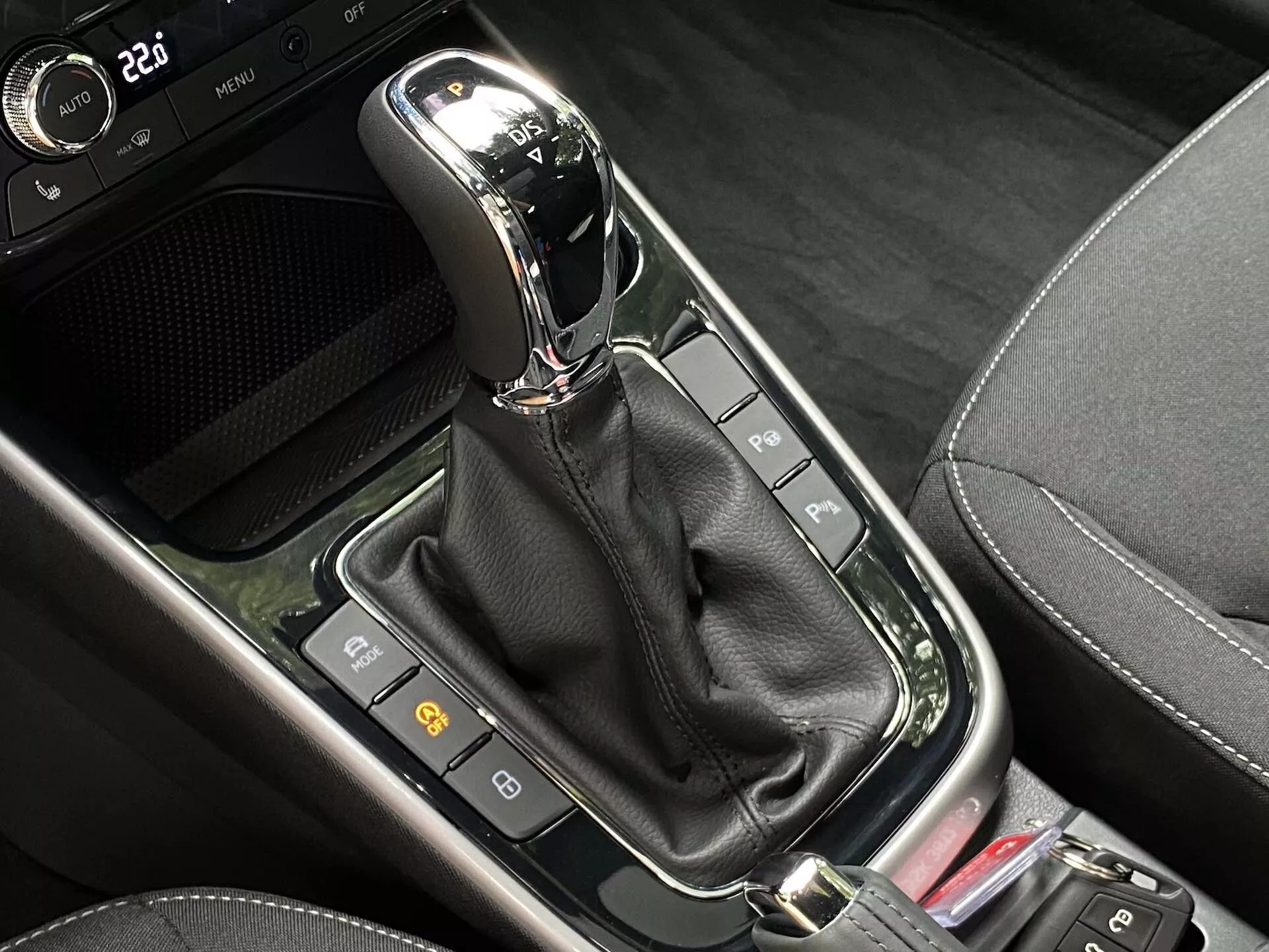 Vista aproximada do seletor de mudanças automático no console central de um Skoda Fabia 2021. O moderno seletor de mudanças, posicionado verticalmente em modo 'P' (Parquear), apresenta um acabamento cromado ou metálico na parte superior e está encapsulado em um painel preto brilhante. Abaixo dele, uma bota de couro preto oculta o mecanismo. Ao redor do seletor de mudanças há uma série de botões e indicadores, incluindo dois à esquerda provavelmente relacionados a funções de segurança do carro, e parte de um botão vermelho à direita. A área combina diversos materiais pretos, incluindo superfícies brilhantes e padrões que imitam fibra de carbono, sugerindo um design contemporâneo de alta tecnologia.