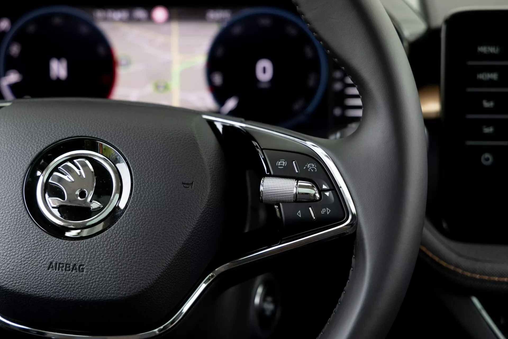Vista detalhada do interior do Skoda Fabia 2021 a partir da perspectiva do motorista, focando nos controles do volante direito. O volante revestido em couro apresenta um emblema central prateado com o logotipo Skoda e a palavra 'AIRBAG' em baixo relevo abaixo. O grupo de controle à direita inclui botões para o sistema de infotainment, chamadas em viva-voz ou comandos de voz, com borboletas visíveis atrás do volante. Fundo desfocado mostra o painel de instrumentos com tacômetro e velocímetro, além de um display digital central para assistência ao motorista. Consola central ou sistema multimédia parcialmente visível à direita.