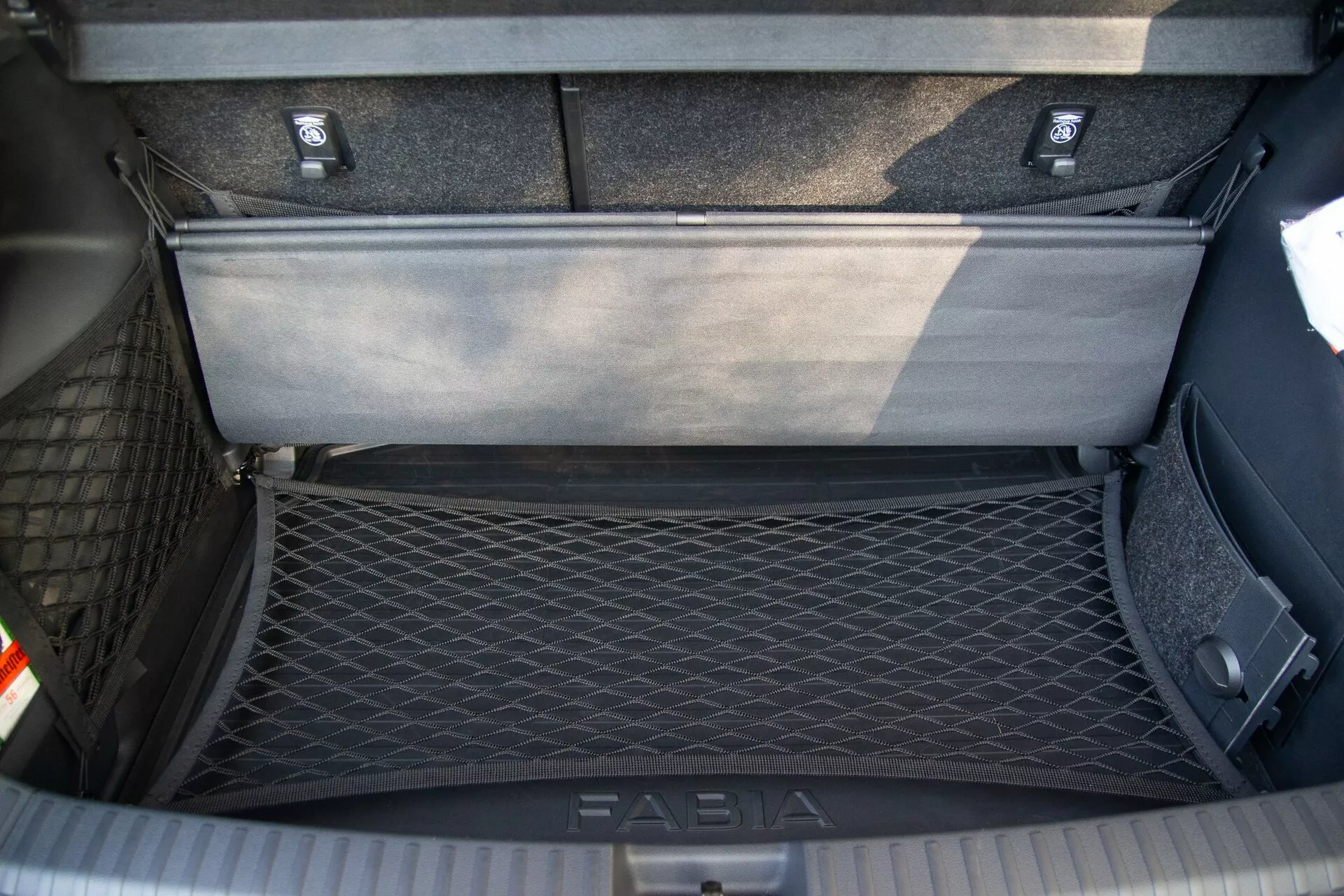 Vista de cima da área do porta-malas de um Skoda Fabia 2021, mostrando o compartimento de armazenamento interno detalhado com uma rede de carga no lado esquerdo para fixação de objetos e um painel de chão flexível parcialmente levantado revelando uma área de armazenamento adicional abaixo. O porta-malas apresenta carpete durável e uma placa de soleira com a marca 'FABIA' para proteção durante o carregamento e descarregamento.