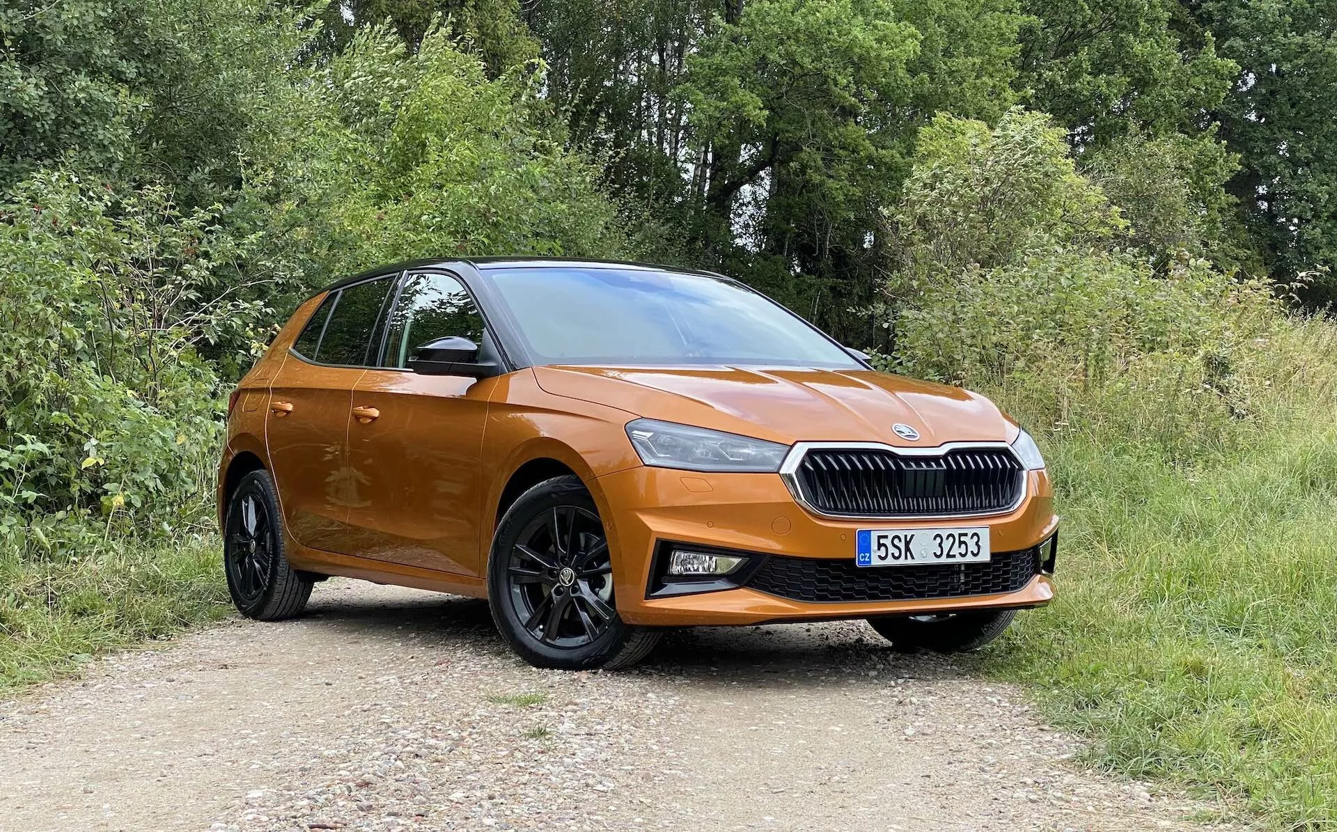 Perfil lateral completo de um moderno hatchback Skoda Fabia 2021 cor de laranja estacionado em um caminho não pavimentado com vegetação ao fundo, capturado em um ângulo ligeiramente oblíquo da frente, destacando o lado do motorista. Características incluem uma grade proeminente com o emblema da marca central, faróis elegantes, luzes de neblina, grandes rodas de liga leve preto de múltiplos raios, maçanetas e espelhos na cor da carroceria, linha do teto inclinada e uma sugestão da traseira e conjunto de luzes.