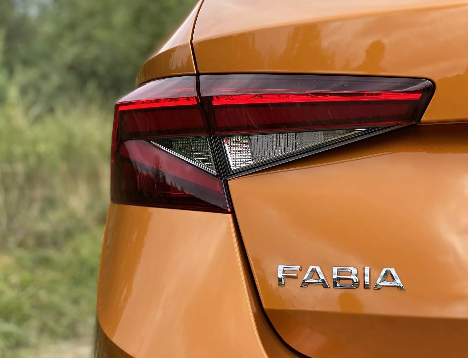 Imagem em close-up do canto traseiro de um Skoda Fabia 2021 destacando o design angular e afiado do conjunto de luzes traseiras de LED e a distinta marca 'FABIA' na carroceria de tom metálico laranja brilhante. As curvas suaves e as pequenas saliências ao redor das luzes traseiras aumentam a estética moderna, capturada num ângulo ligeiramente elevado e com pouca profundidade de campo.