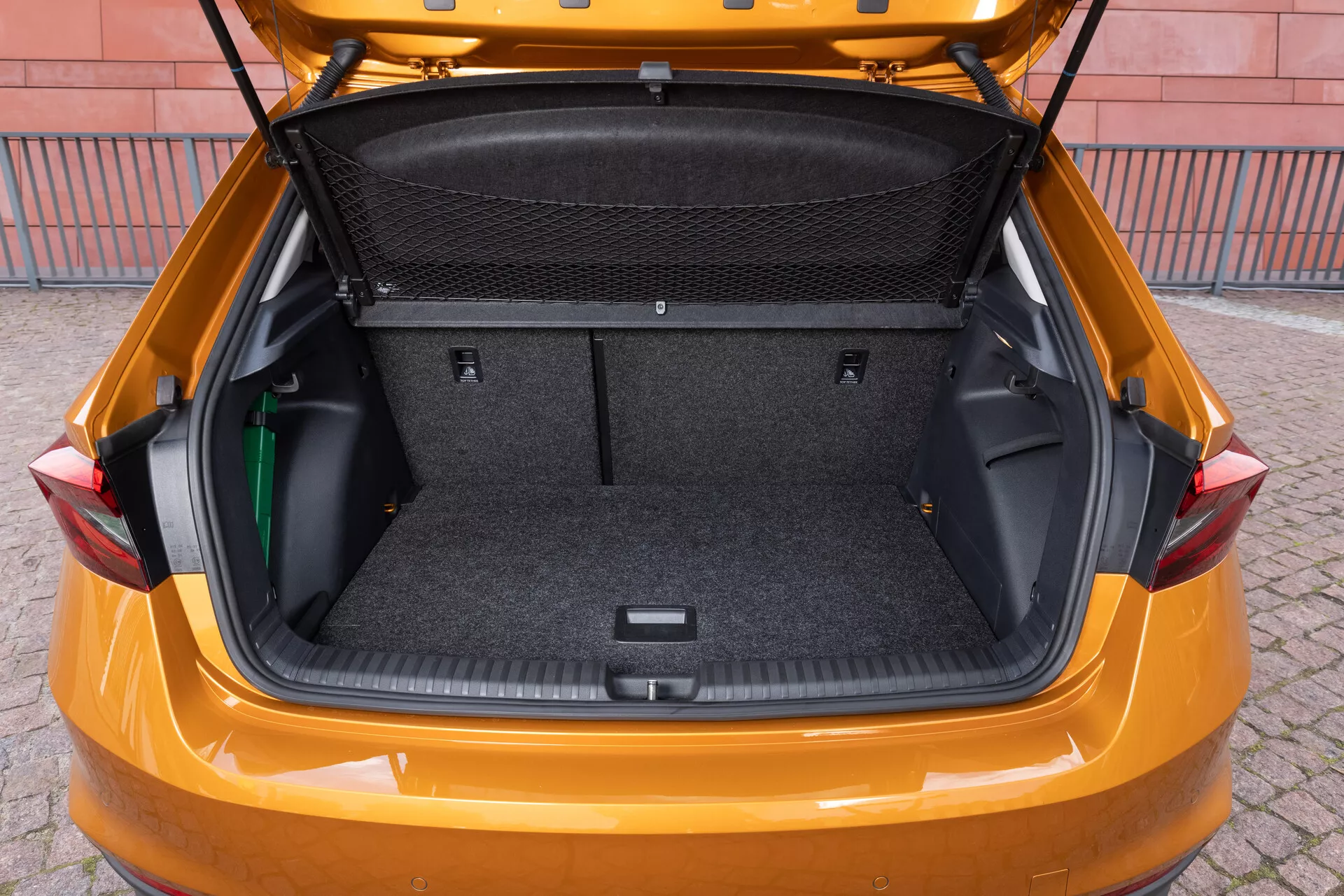 A imagem retrata o porta-malas de um modelo Skoda Fabia 2021, fotografado de um ângulo traseiro ligeiramente elevado para mostrar o espaçoso compartimento de carga. O interior é revestido com um material tipo carpete e possui áreas de armazenamento laterais flexíveis, uma divisão em rede para a carga e uma área rebaixada na parte inferior para armazenamento adicional. A porta traseira está levantada, revelando um porta-malas grande e limpo, projetado para destacar a capacidade de armazenamento e a praticidade do veículo.