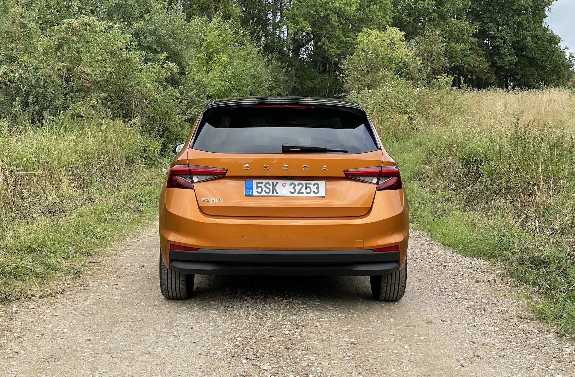 Vista traseira de um hatchback Skoda Fabia 2021 laranja metálico brilhante fotografado à luz do dia em um caminho estreito de terra em um ambiente rural. A imagem captura o design traseiro moderno com uma barra de luz traseira contínua, luzes traseiras angulares, refletores integrados, limpador de para-brisa traseiro e spoiler traseiro para aerodinâmica, destacando o acabamento brilhante e os recursos práticos do carro.