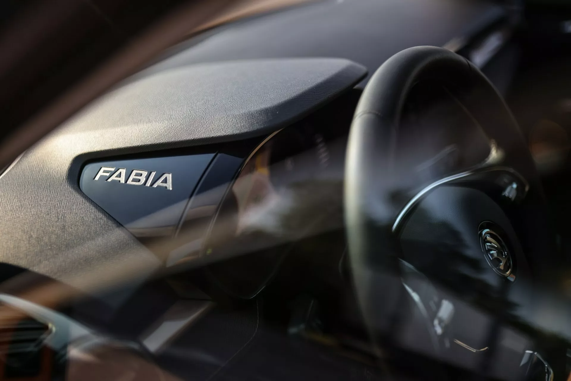 Vista interna em close-up do Skoda Fabia 2021, destacando o lado do passageiro do painel. A palavra 'FABIA' é exibida com destaque, enquanto o volante moderno com o logotipo Skoda é parcialmente visível à esquerda. O painel apresenta uma textura granulada e a pequena profundidade de campo enfatiza a marca com um fundo ligeiramente desfocado.