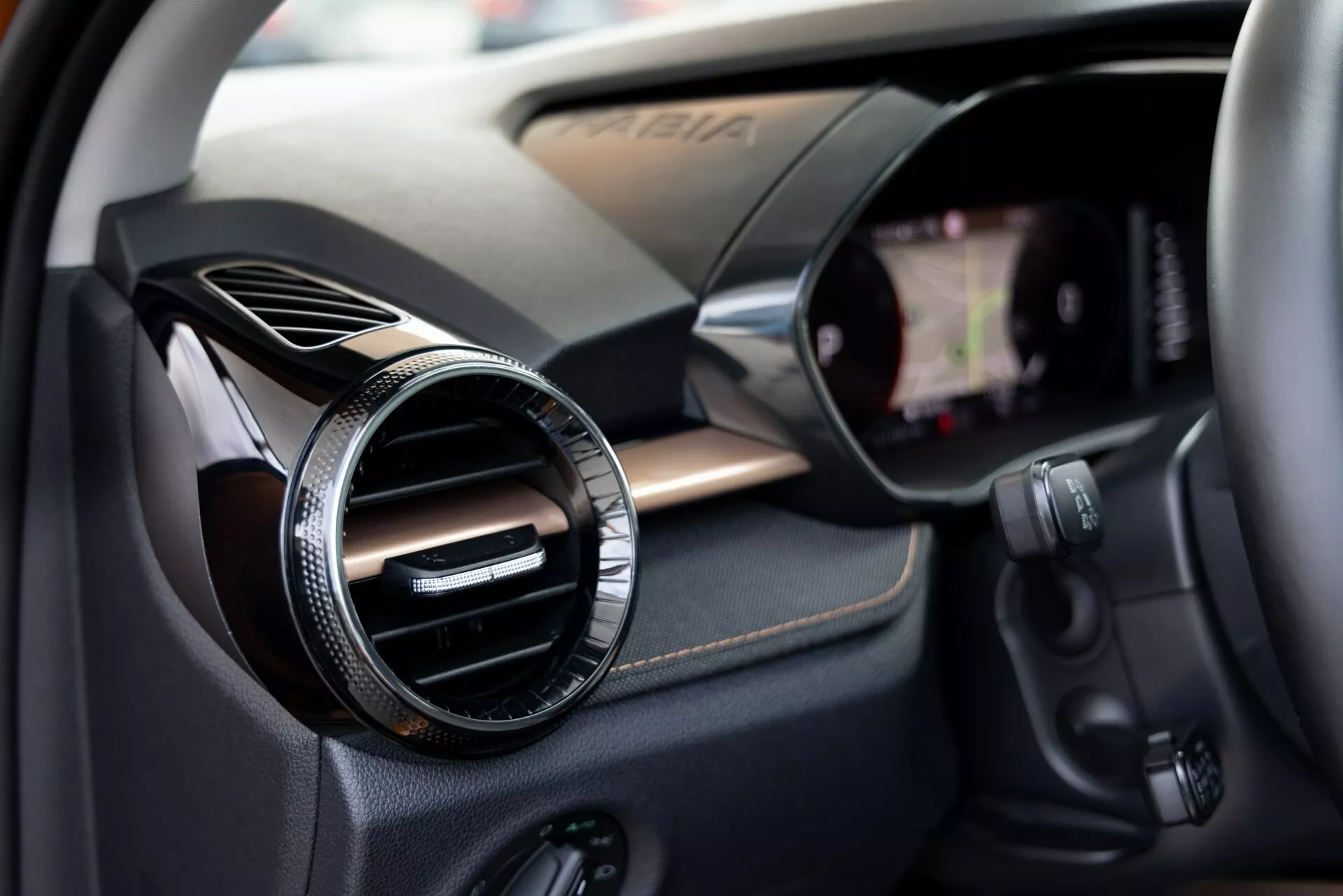 Close-up do interior do Škoda Fabia 2021 focando no lado do passageiro. A imagem captura uma ventilação redonda com acabamento metálico ou cromado contra um painel texturizado semelhante a fibra de carbono, indicando um acabamento esportivo ou de alta qualidade. À direita está visível a borda de uma tela touchscreen de infotainment integrada no painel. Abaixo da ventilação estão vários botões de controle, possivelmente para o sistema de áudio, controle de clima ou outras funcionalidades do veículo. A pequena profundidade de campo chama a atenção para a ventilação enquanto suavemente sugere os componentes tecnológicos ao redor.