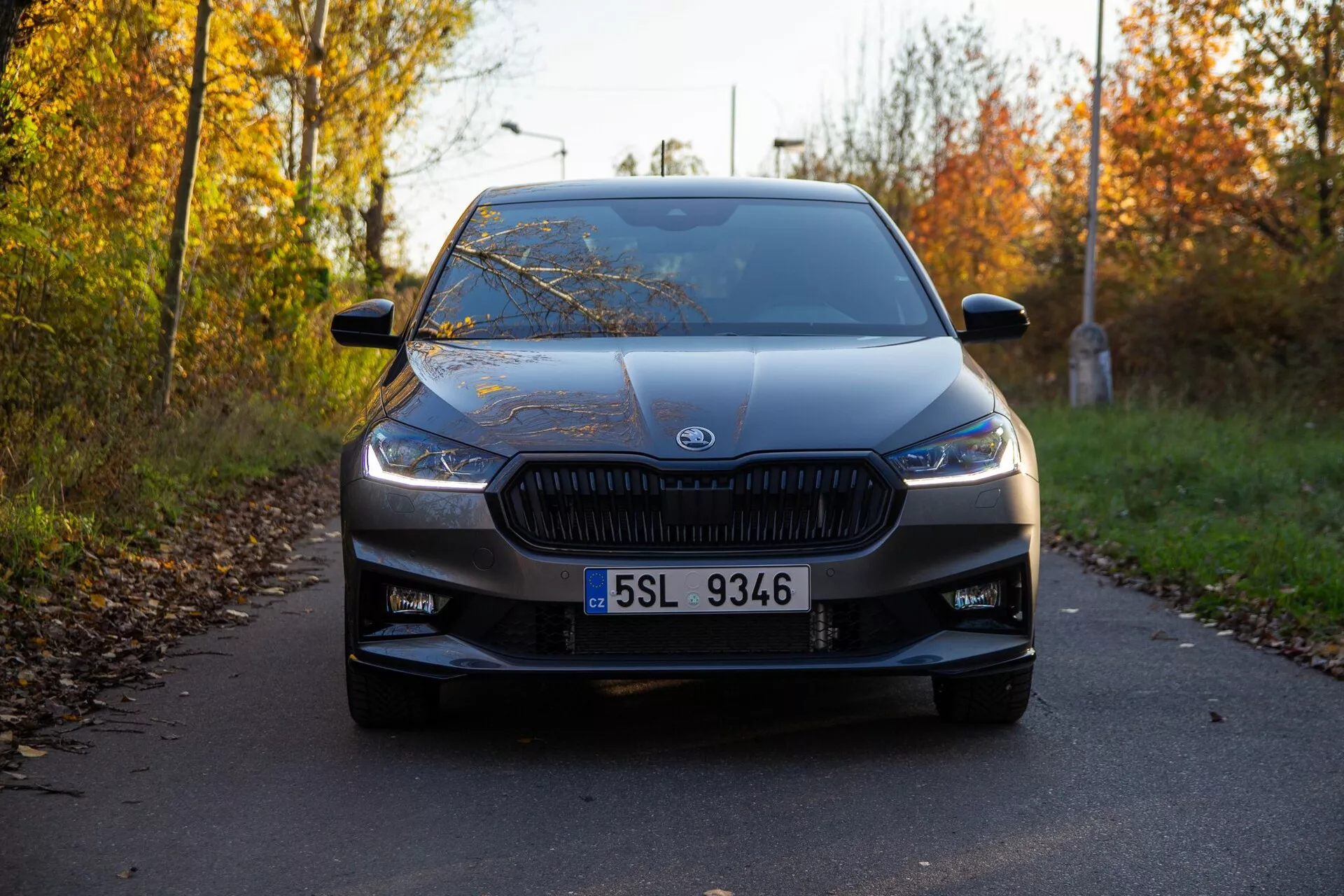 Vista frontal de um Skoda Fabia 2021 capturada de um ângulo baixo, destacando sua grade distintiva com lamelas verticais e faróis modernos e estreitos. O carro, centralizado na imagem, está estacionado em uma estrada estreita ladeada por folhagem de outono, com o emblema do fabricante proeminente no centro da grade. Outros detalhes de design visíveis incluem a placa de licença clara, entradas de ar no para-choque inferior, linhas de contorno suaves no capô, para-brisa expansivo e retrovisores laterais com indicadores. A iluminação suave e as sombras longas sugerem que a imagem foi tirada no início da manhã ou no final da tarde, mostrando o carro em excelente estado.