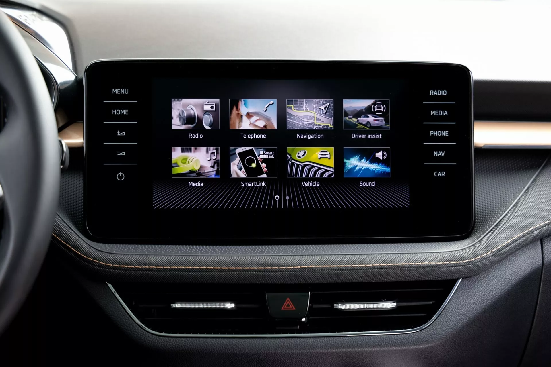 Vista aproximada do sistema de infoentretenimento com tela sensível ao toque do Škoda Fabia 2021, a partir da perspectiva de um passageiro ou motorista no assento dianteiro, mostrando um console central elegante, emoldurado possivelmente por alumínio escovado. A tela exibe uma interface gráfica amigável com ícones coloridos para Home, Rádio, Telefone, Navegação e mais. Abaixo da tela, uma fileira de botões físicos e uma possível fenda para CD adicionam multifuncionalidade. O revestimento premium com costura contrastante sugere um design interior de alta qualidade.