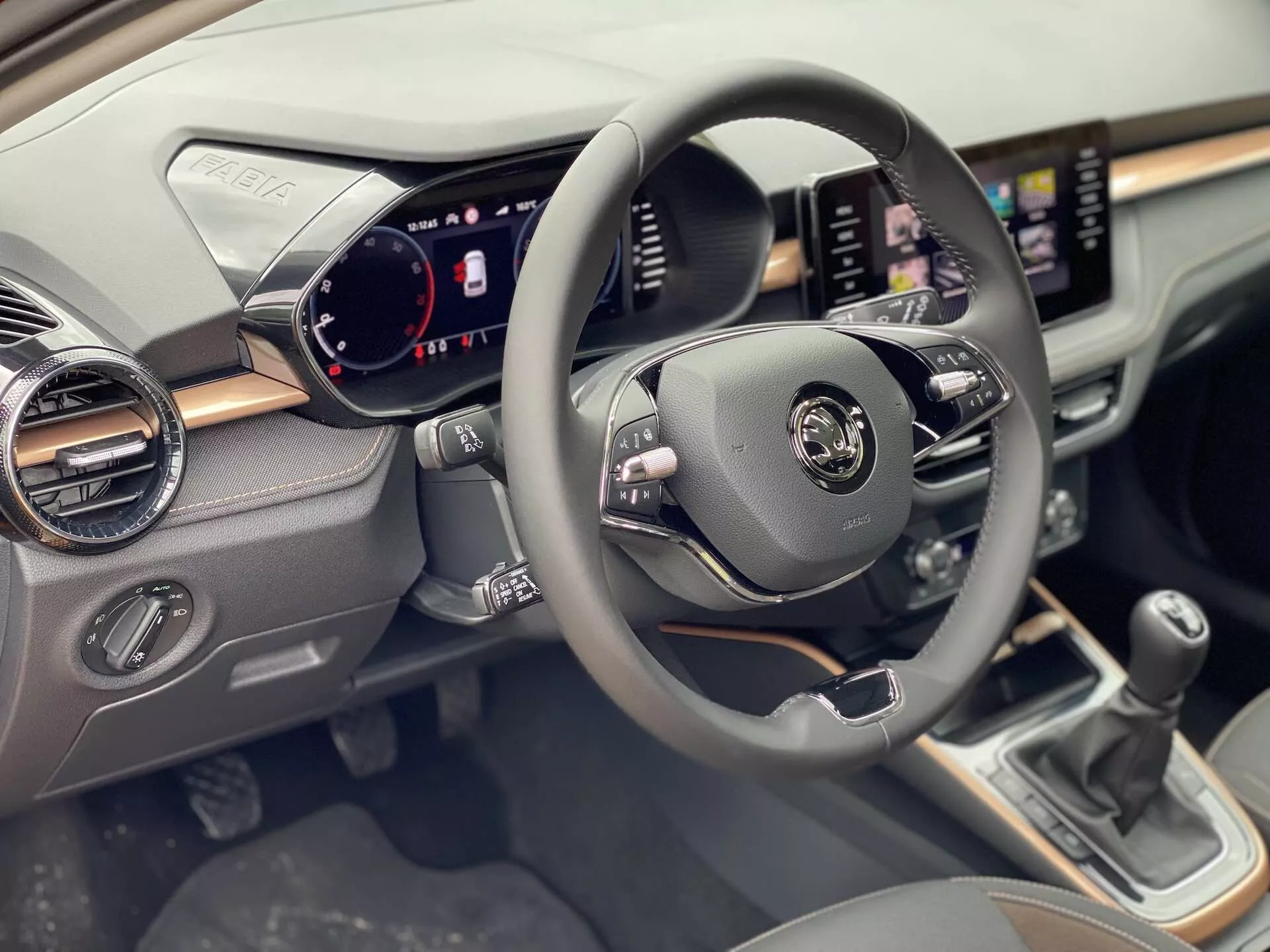 Vista interior do Skoda Fabia 2021 focada no lado do condutor, mostrando o volante centralizado com controles multifuncionais para áudio, comandos de voz e controle de cruzeiro. O painel de instrumentos digital exibe velocímetro, tacômetro e indicadores de status do veículo. Um sistema de infotainment com tela sensível ao toque é exibido no painel com funcionalidades de navegação e mídia. Abaixo da tela, há botões e controles do sistema de climatização. A alavanca de câmbio manual é visível no console central.