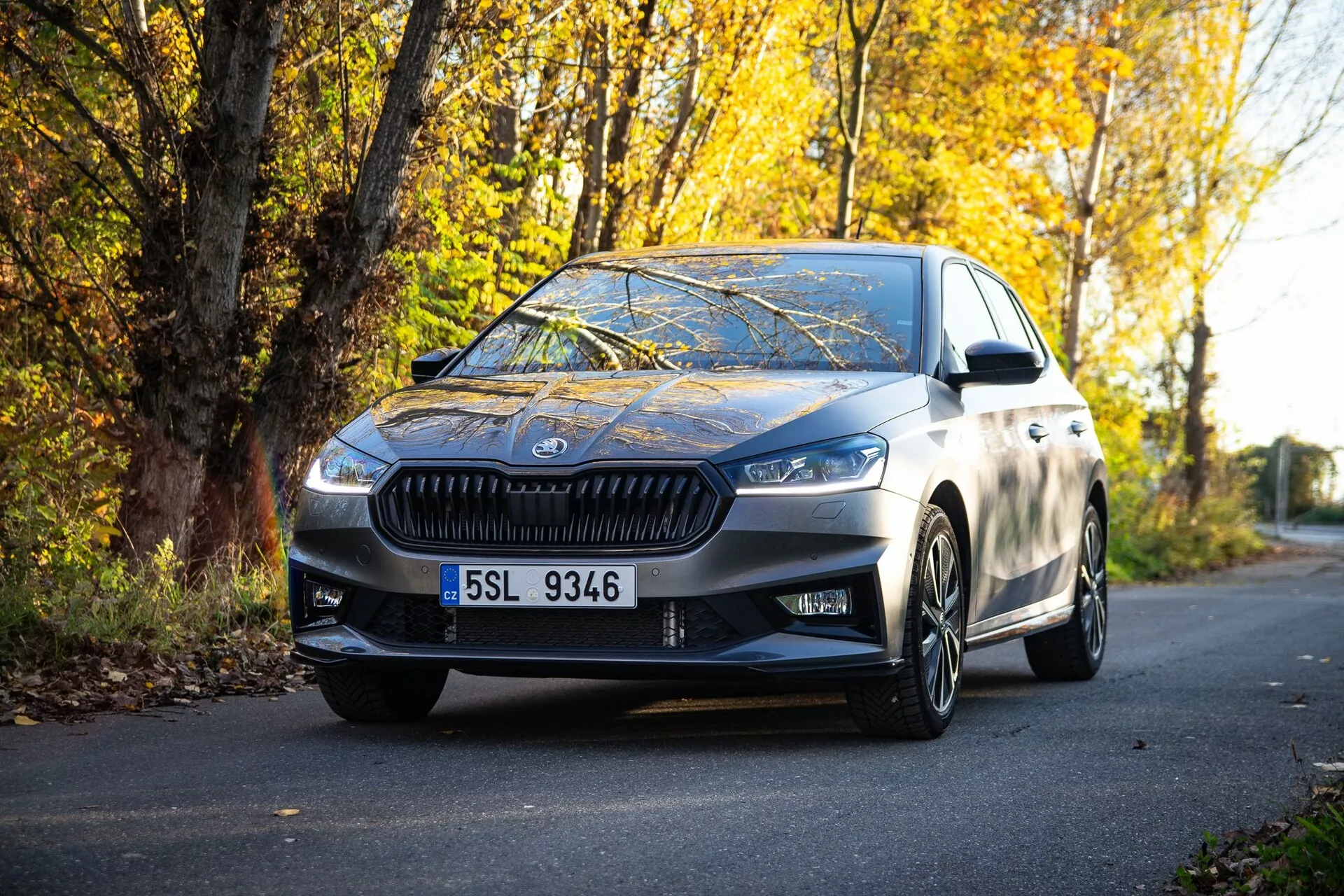 Imagem de um sedan Skoda Fabia 2021 moderno estacionado em uma estrada ladeada por folhagem de outono, capturada de uma perspectiva ao nível do solo, destacando a grade frontal, faróis angulares, linhas de design elegantes, rodas de liga leve e espelhos laterais com indicadores de direção integrados, refletindo um acabamento metálico de alta qualidade e uma mistura de luxo e esportividade.