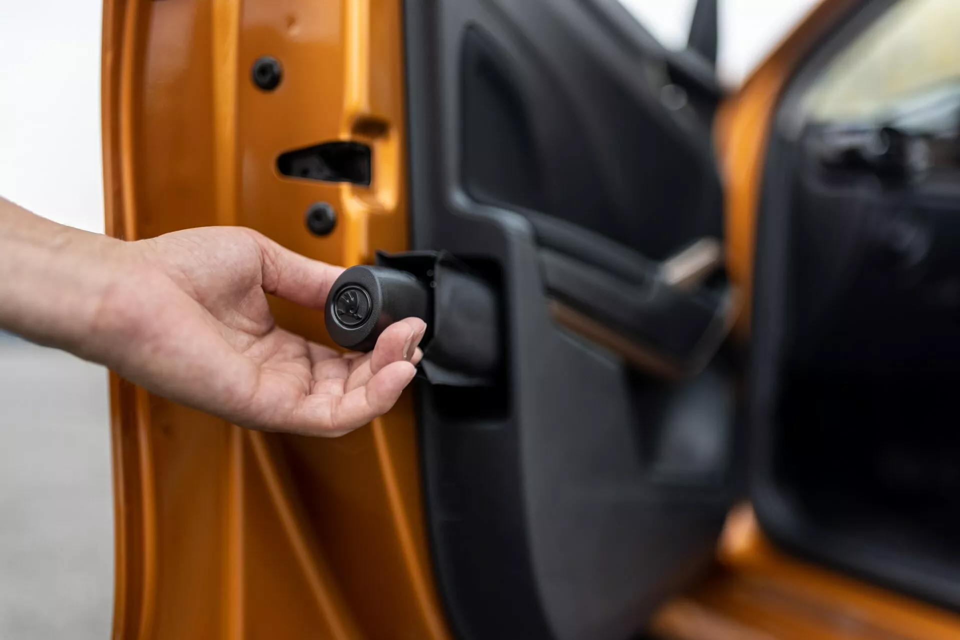 Close-up da porta do motorista do Skoda Fabia 2021, ligeiramente aberta, vista do exterior com a borda interior visível. Uma mão segura um chaveiro compacto, preto e circular, que indica um sistema de entrada sem chave ou sistema de segurança automotiva. O carro é laranja brilhante com um acabamento metálico, apresentando mecanismos de bloqueio avançados e recursos de segurança personalizados.