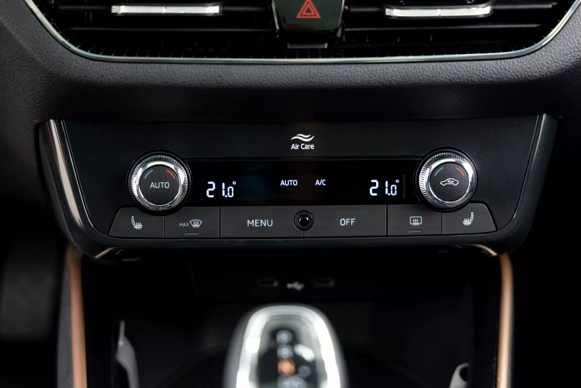 Vista de perto do painel de controle climático do Skoda Fabia 2021 localizado no console central, com controles de temperatura de dupla zona com visores digitais mostrando '20', botões integrados para controle climático automático ('AUTO'), ar condicionado ('AC'), configurações de menu ('MENU') e desligamento do sistema ('OFF'). Uma moldura preta brilhante envolve uma tela de informações acima dos controles, com um botão de aviso de perigo (ícone de triângulo) visível no topo. O painel está inserido em um interior premium com acabamento fosco e imitação de madeira, destacando um design e tecnologia modernos.