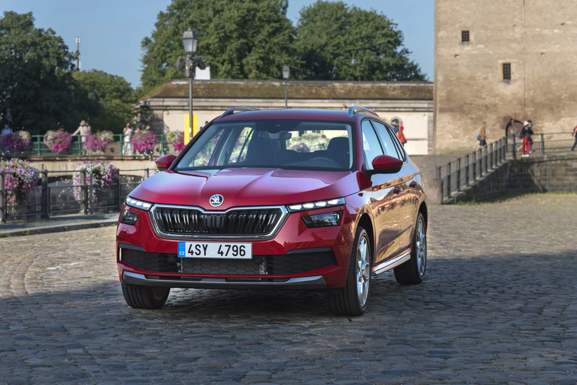 Skoda Kamiq 2019 em vermelho, fotografado de um ângulo frontal de três quartos, destacando a grade frontal, faróis LED, capô, para-choque dianteiro com possíveis luzes de neblina ou entradas de ar, e rodas de liga leve, contra um fundo de rua de paralelepípedos urbana ou histórica com folhagem.