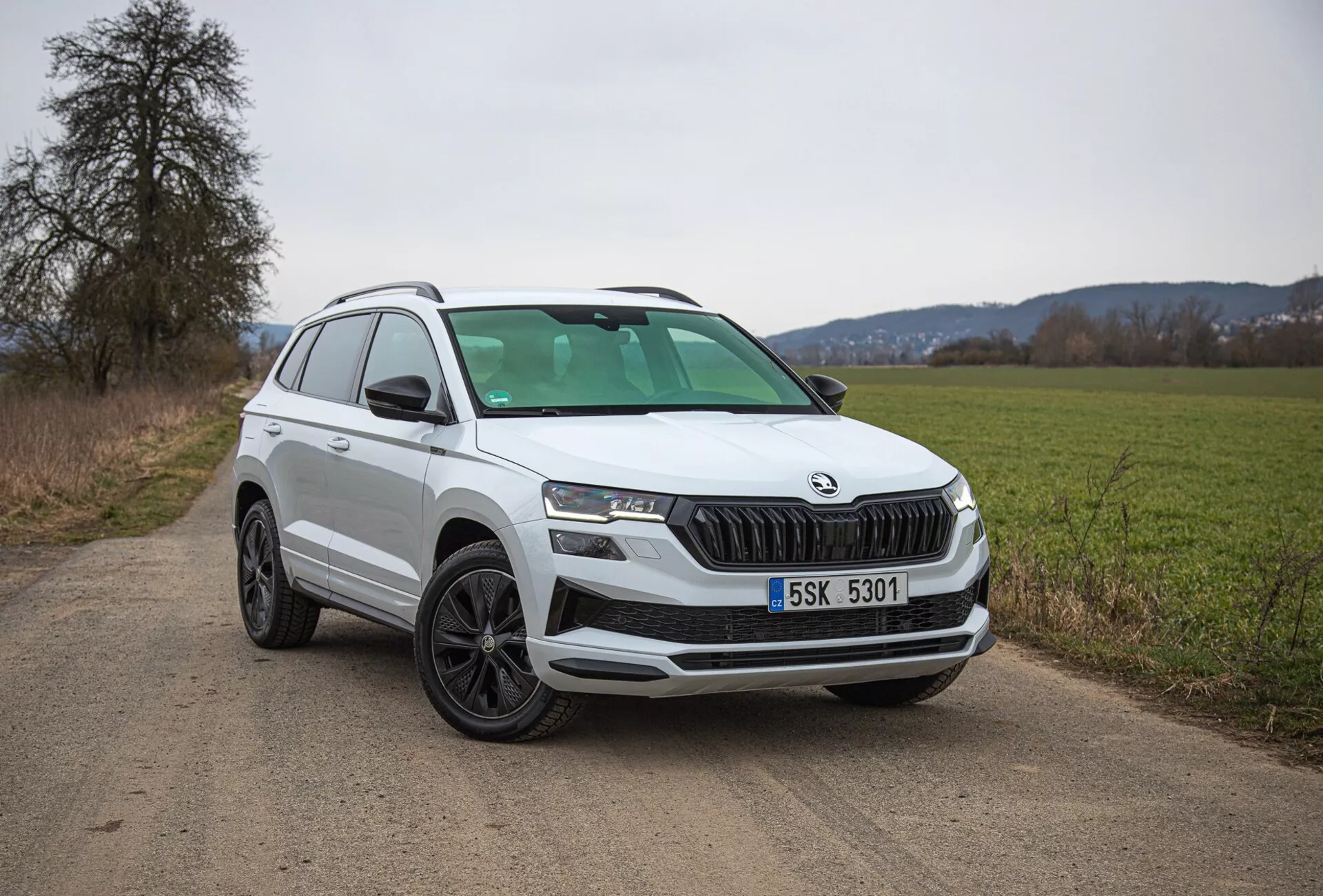 SUV branco Škoda Karoq 2021 estacionado em uma estrada de terra rural, mostrando a vista do quarto frontal esquerdo incluindo grade, faróis e design de roda, com foco em características modernas como iluminação LED e sensores de estacionamento.