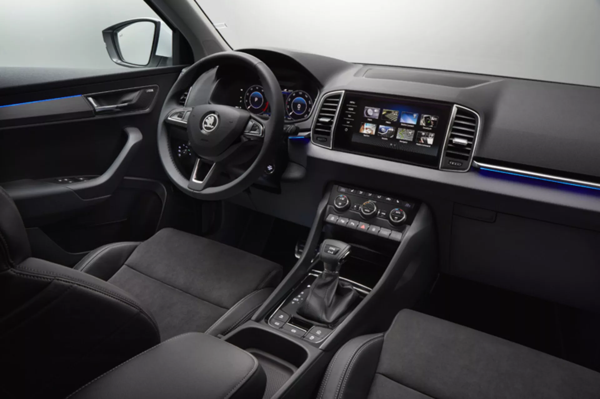 Vista do interior do Skoda Karoq 2017 mostrando o painel de instrumentos, porta do motorista com controles de janelas e espelhos, volante com botões de multimídia e controle de cruzeiro, cluster de instrumentos digital-analógico, sistema de infotenimento touchscreen, controle de clima de zona dupla, seletor de marcha automático, seleção de modo de condução e bancos revestidos em couro