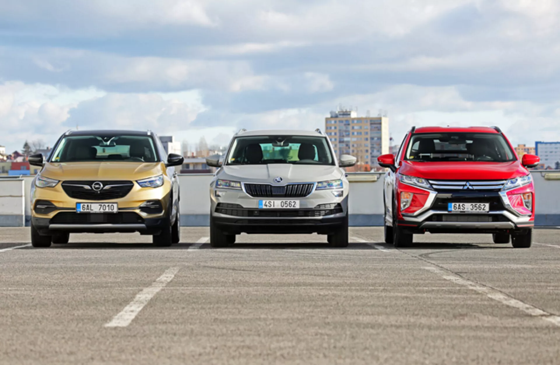Vista frontal de três modelos de Skoda Karoq 2017 em um estacionamento com paisagem urbana ao fundo, mostrando diferentes designs de grade frontal, faróis e formas aerodinâmicas de capô sob condições de luz diurna nublada