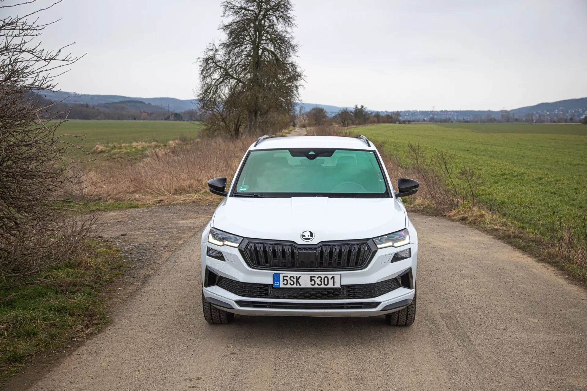 SUV Škoda Karoq 2021 retratando uma visão frontal direta em uma estrada de terra estreita, destacando a grade distintiva da Škoda, faróis de LED afiados, luzes de nevoeiro dentro do contorno preto para capacidade fora de estrada potencial, linhas dinâmicas do capô e recursos tecnológicos avançados na frente do veículo.