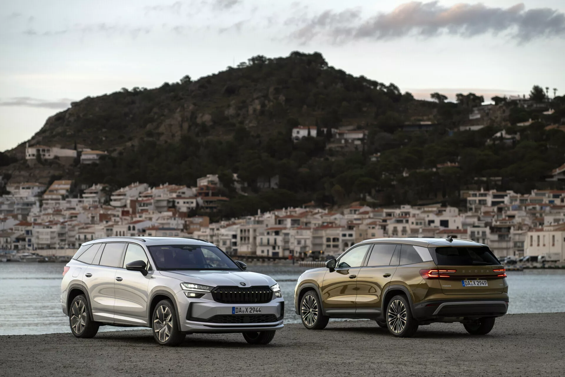 SUVs Skoda Kodiaq 2024 estacionados lado a lado ao amanhecer em um passeio à beira-mar com uma cidade costeira ao fundo, mostrando a grade frontal, faróis e linhas de design elegantes de um e a vista traseira de três quartos do outro.