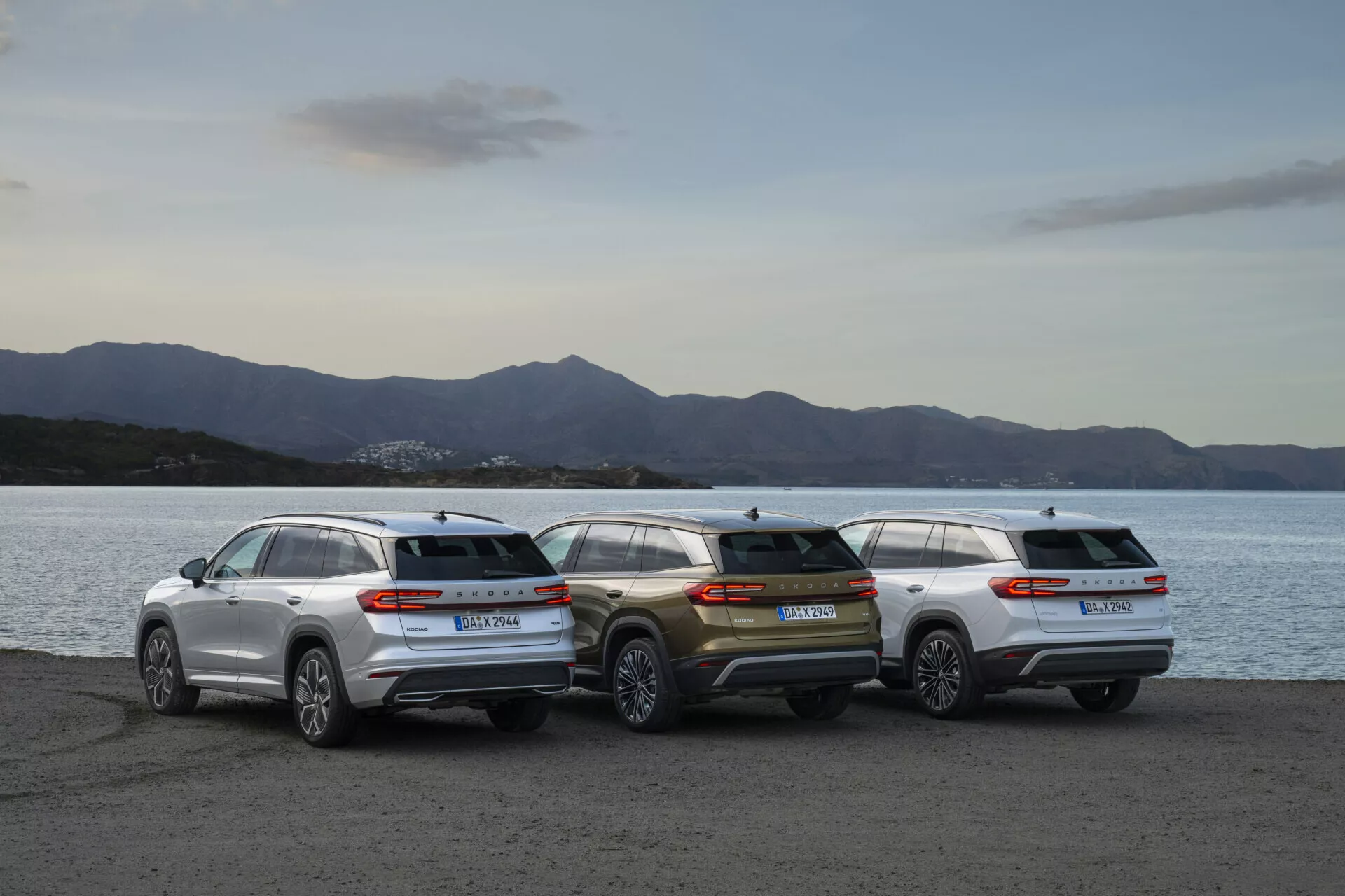 Linha de Skoda Kodiaq 2024 com três SUVs estacionados à beira-mar ao crepúsculo, mostrando perfis traseiros e estética de design contra um fundo de paisagem suavemente iluminado