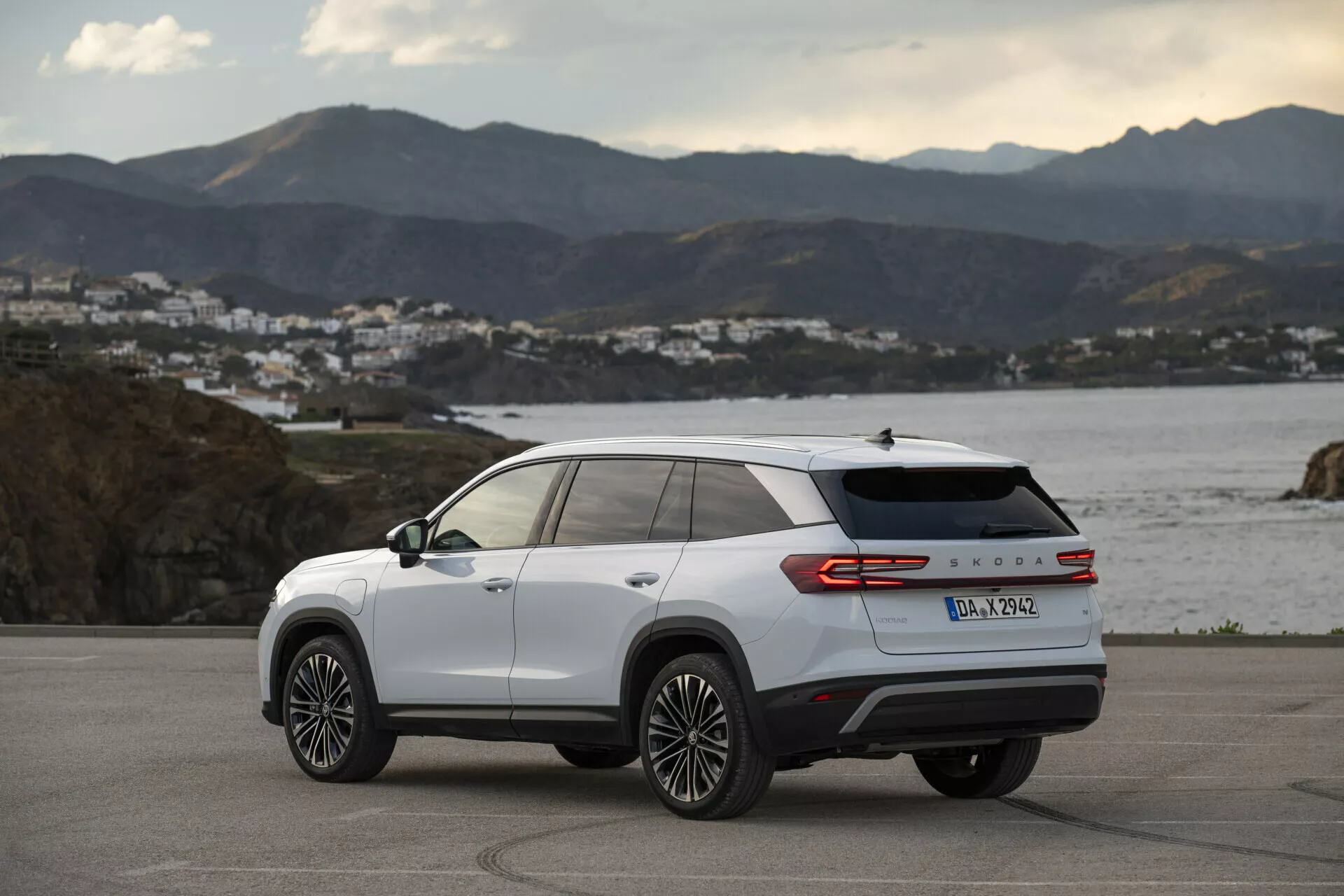 Visão traseira de três quartos de um Skoda Kodiaq 2024 branco, exibindo linhas de design laterais e traseiras, rodas de liga leve multirraios, lanternas traseiras LED e emblema do modelo, estacionado ao ar livre em uma estrada costeira na hora dourada